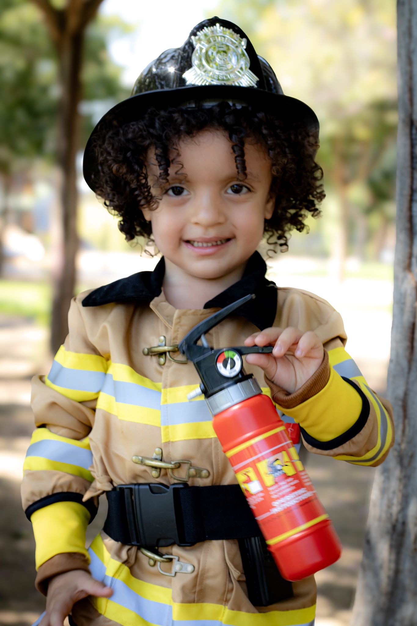 Smiling toddler dressed in a firefighter costume holding a toy fire extinguisher outdoors.