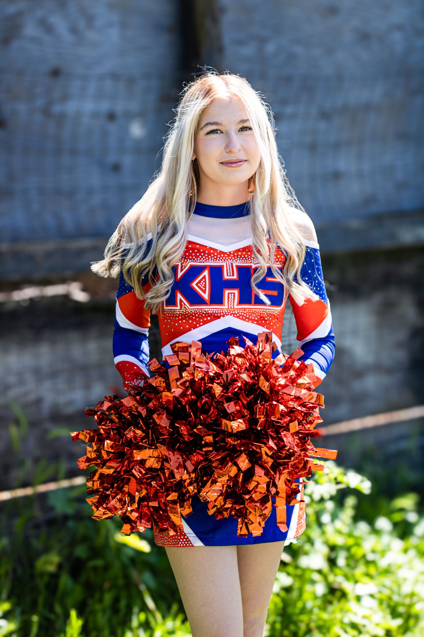 A cheerleader in red, white and blue uniform holds shimmering pom-poms while posing outdoors on a sunny day.