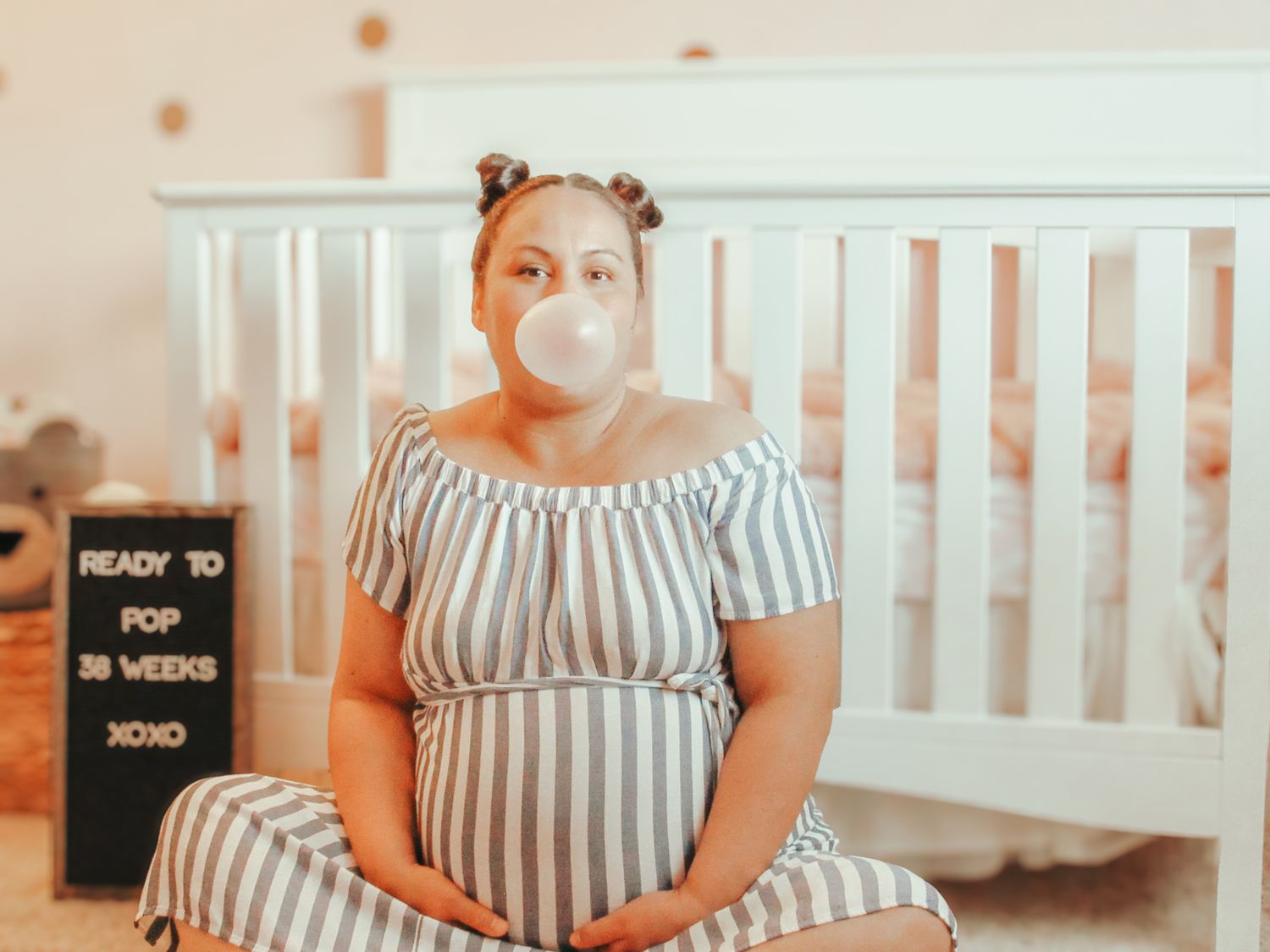 Someone in a striped maternity dress sits on the floor in front of a white crib in a nursery setting.