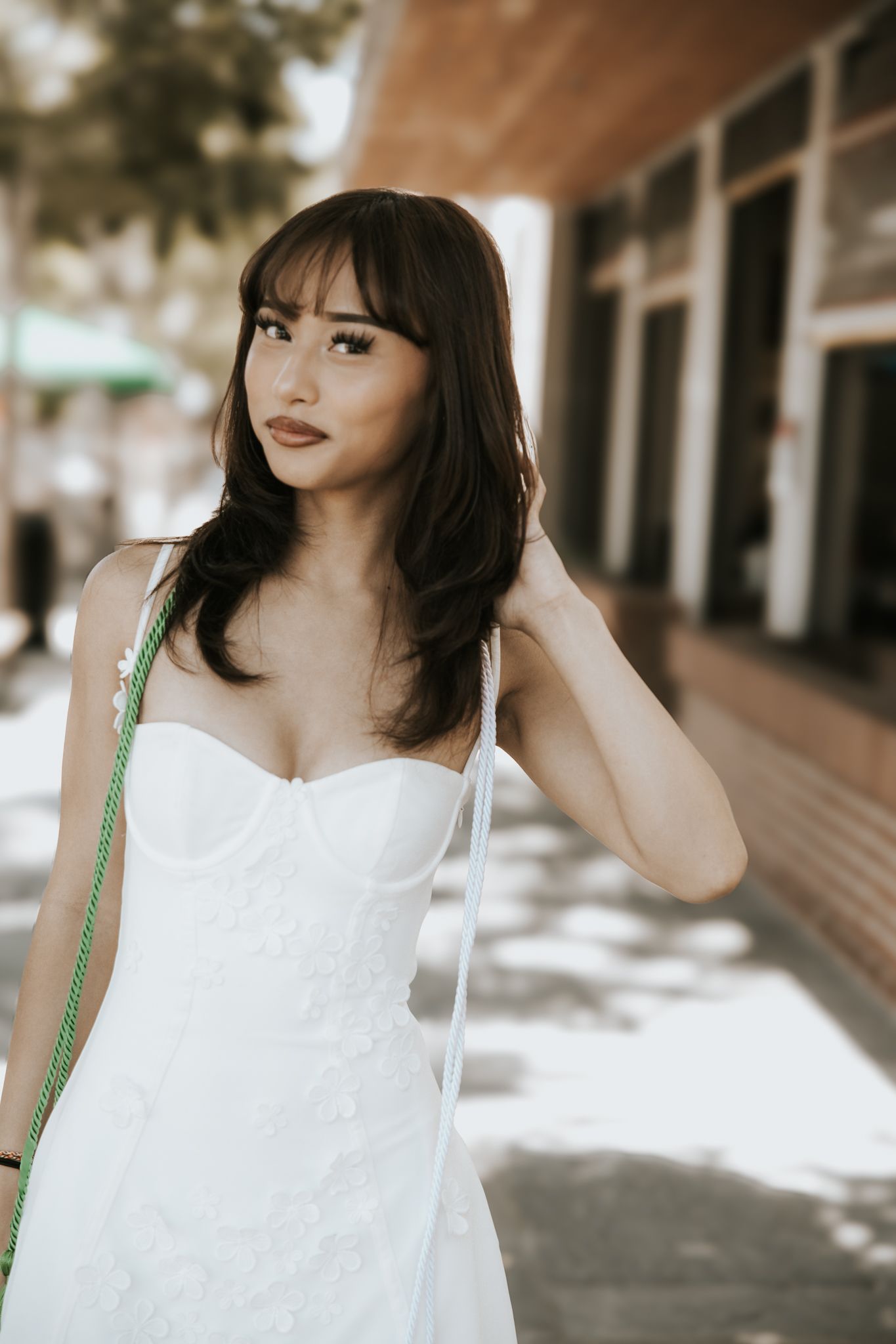 A sophisticated portrait outdoors featuring a sleeveless white dress and green purse strap against a blurred urban backdrop.