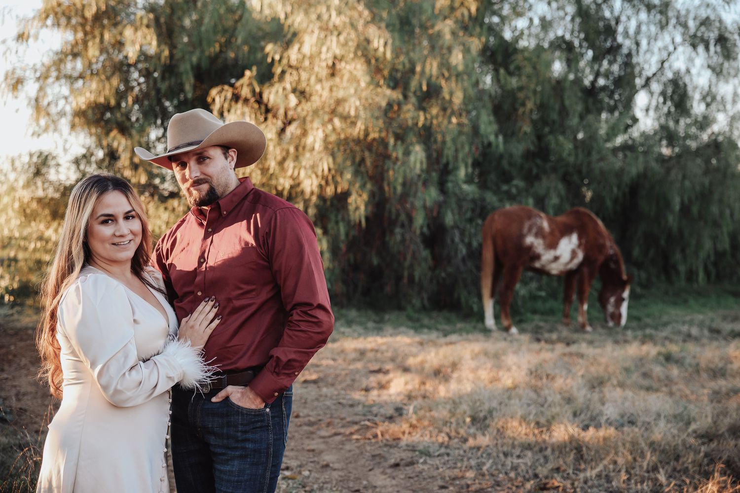 A couple in western attire shares romantic moments during a golden hour photoshoot in a rustic outdoor setting.
