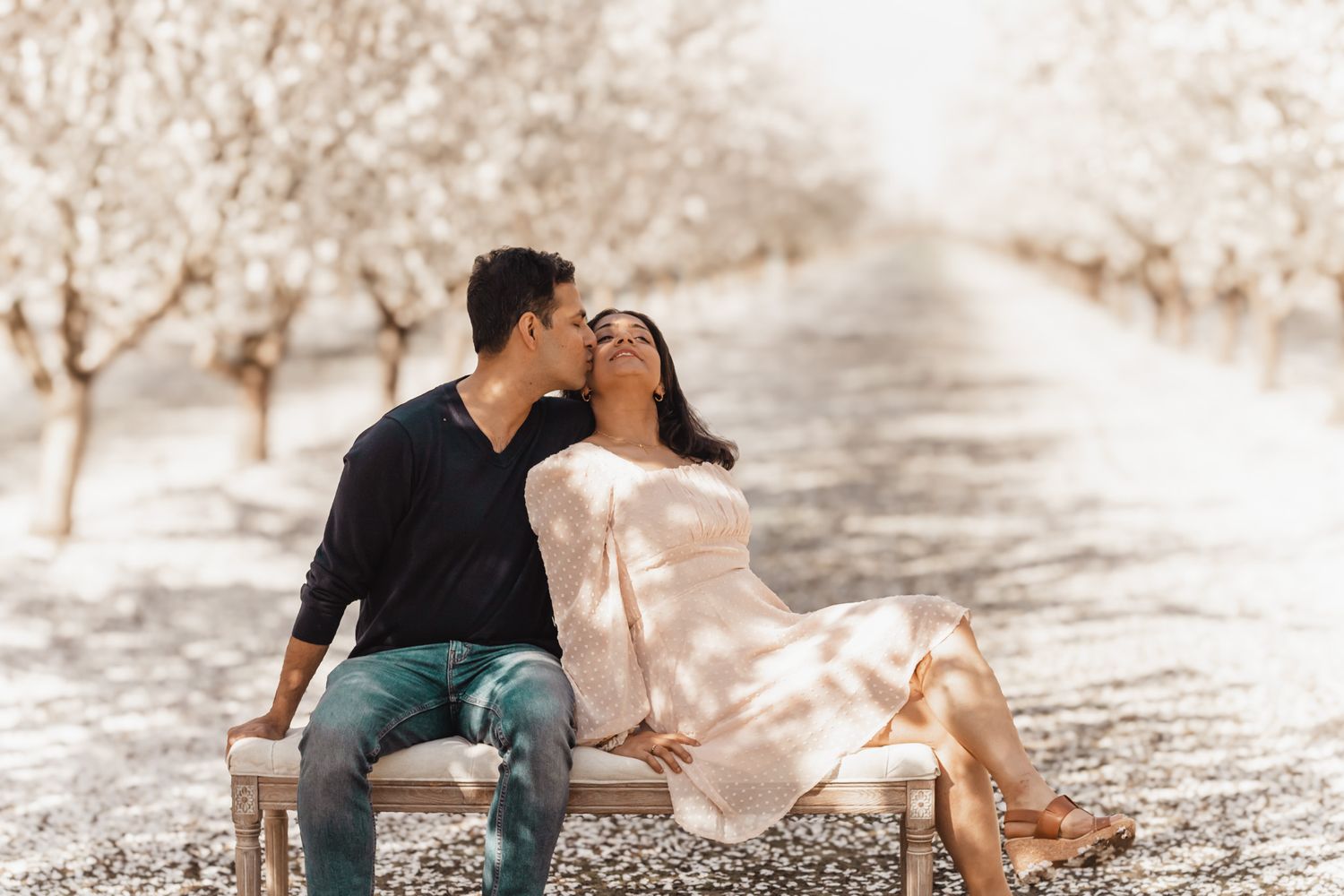 Couple sharing joyful moment on bench along dreamy tree-lined path with soft sepia tones.