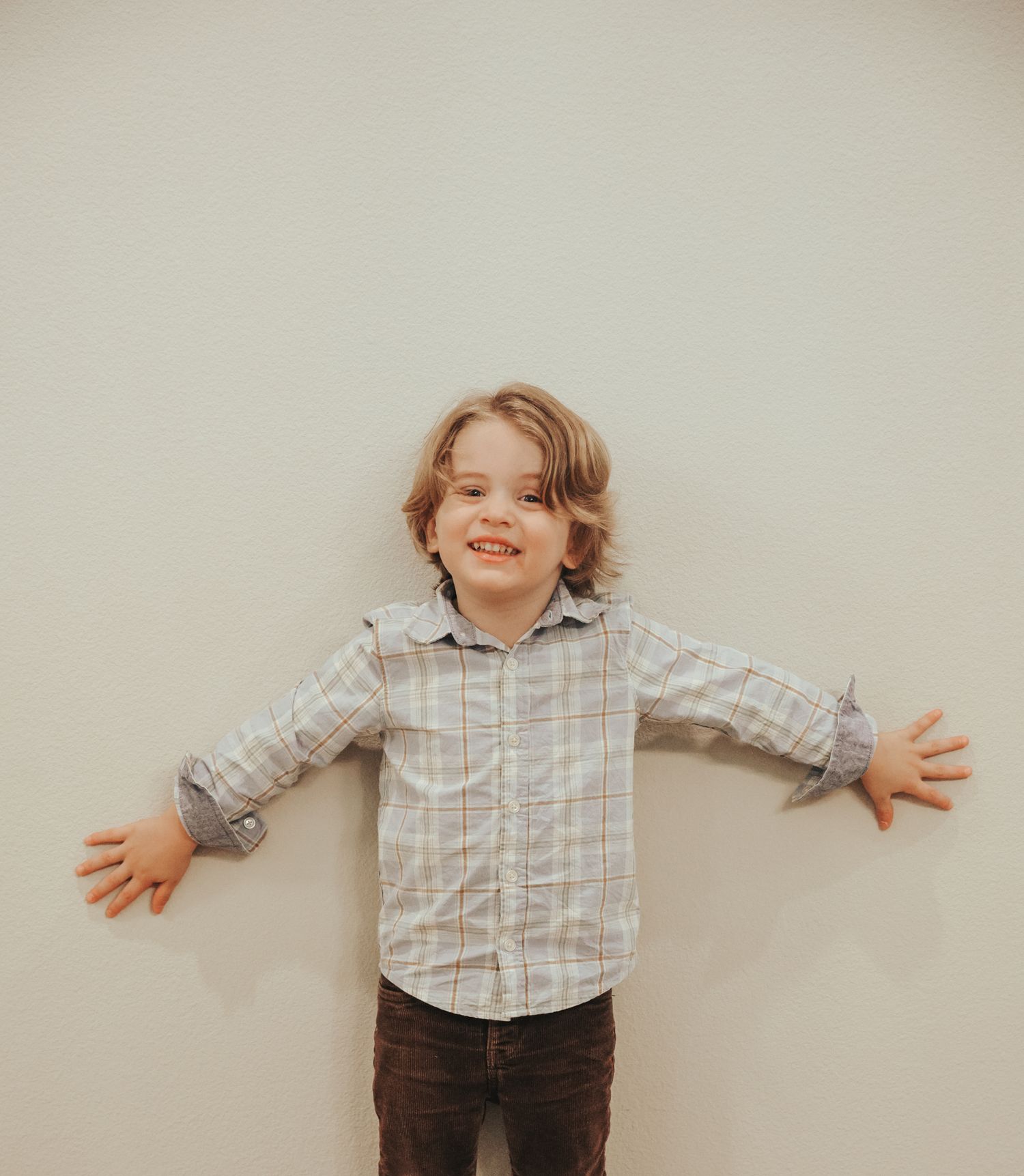 Child in plaid button-down shirt and jeans stands with arms outstretched against neutral background.
