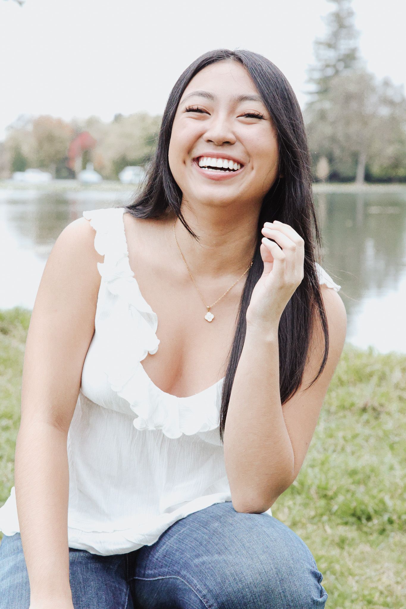 A joyful person in a white sleeveless top sits outdoors near a lake on a sunny day.