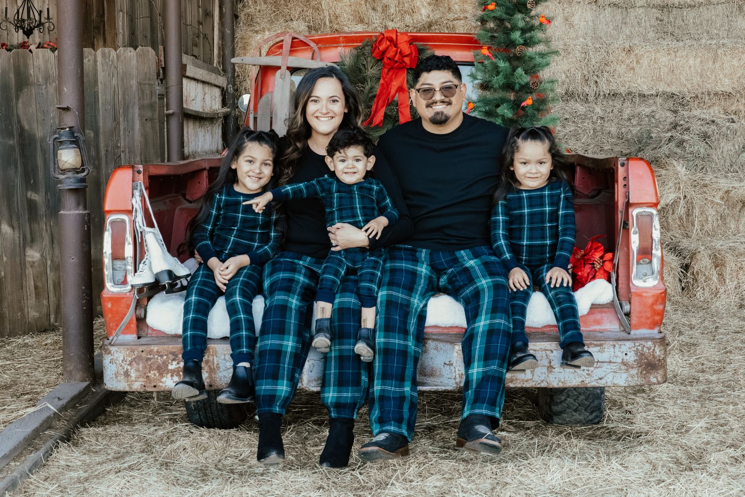 Family in matching blue plaid and black outfits sits together on red vintage truck tailgate with Christmas tree.
