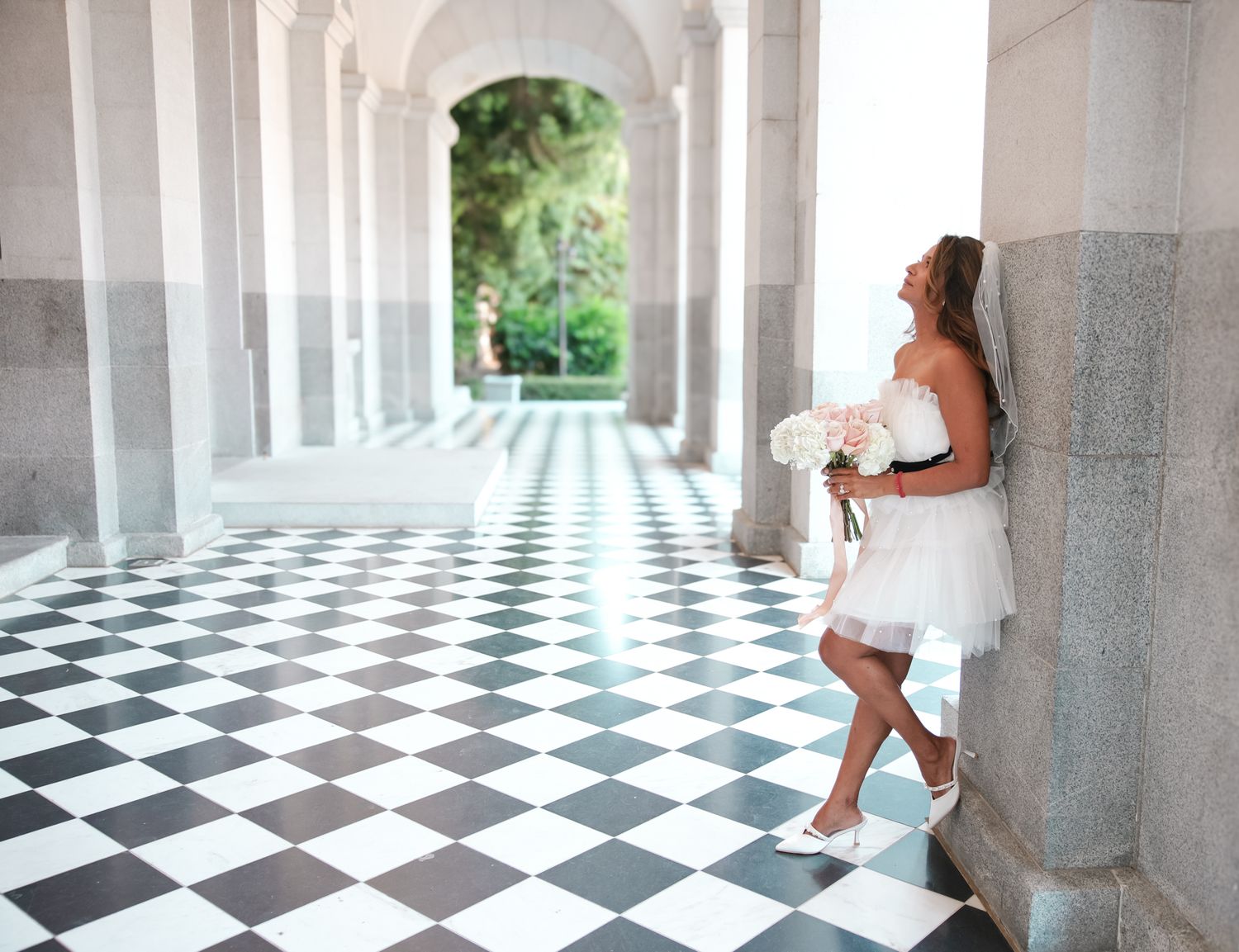 Figure in a white dress poses against a column in an elegant corridor with black and white checkered floor.