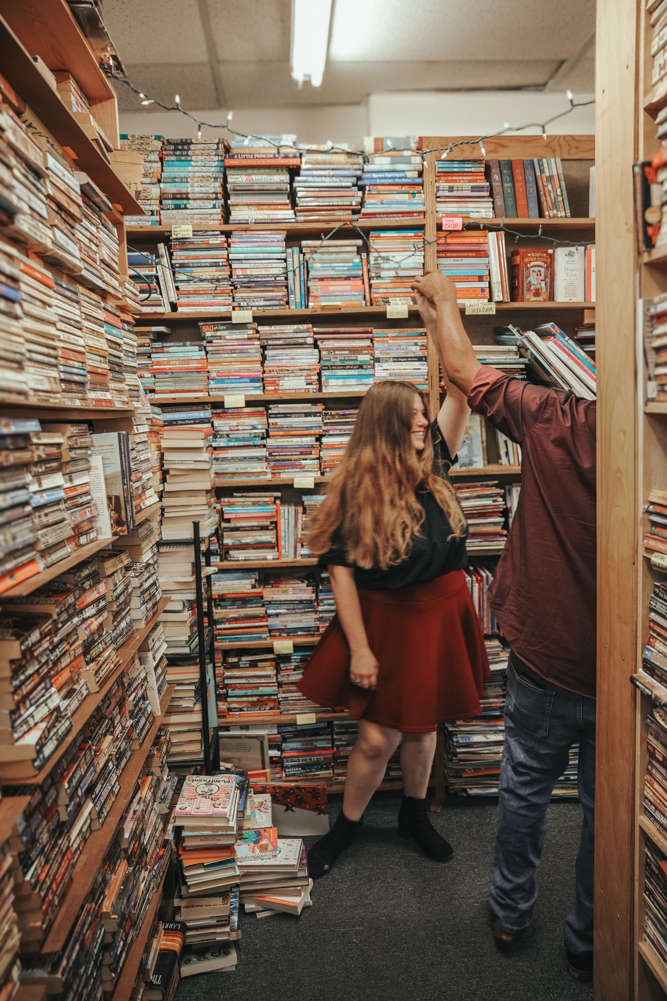 A narrow aisle in a used bookstore is stacked floor to ceiling with piles of books on wooden shelves.