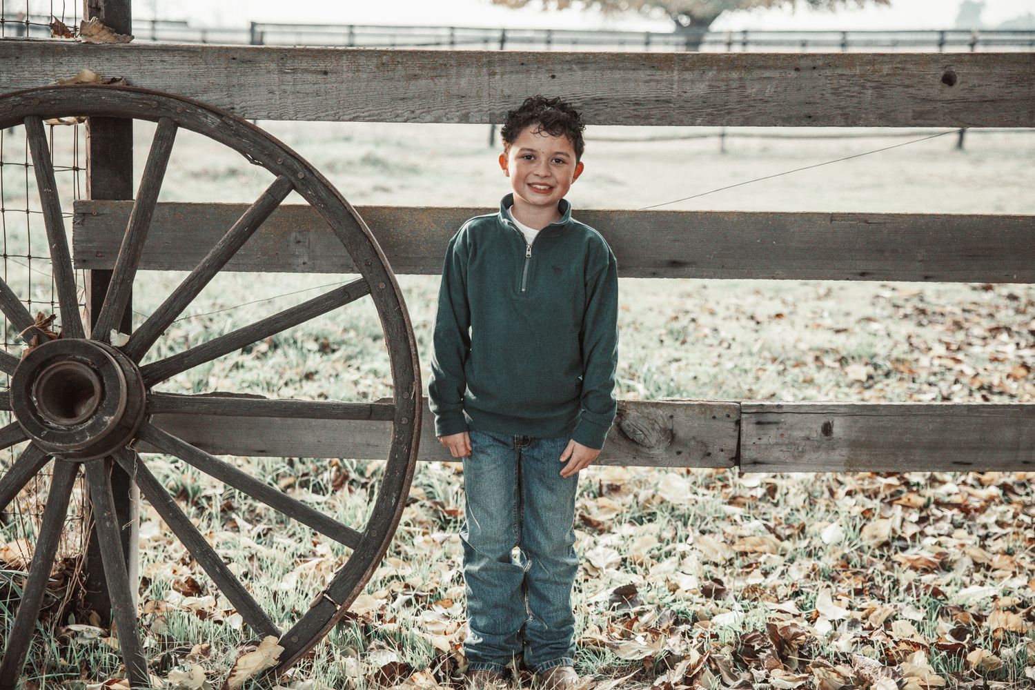 A rustic wooden wagon wheel leans against a white fence with autumn leaves scattered on the ground.