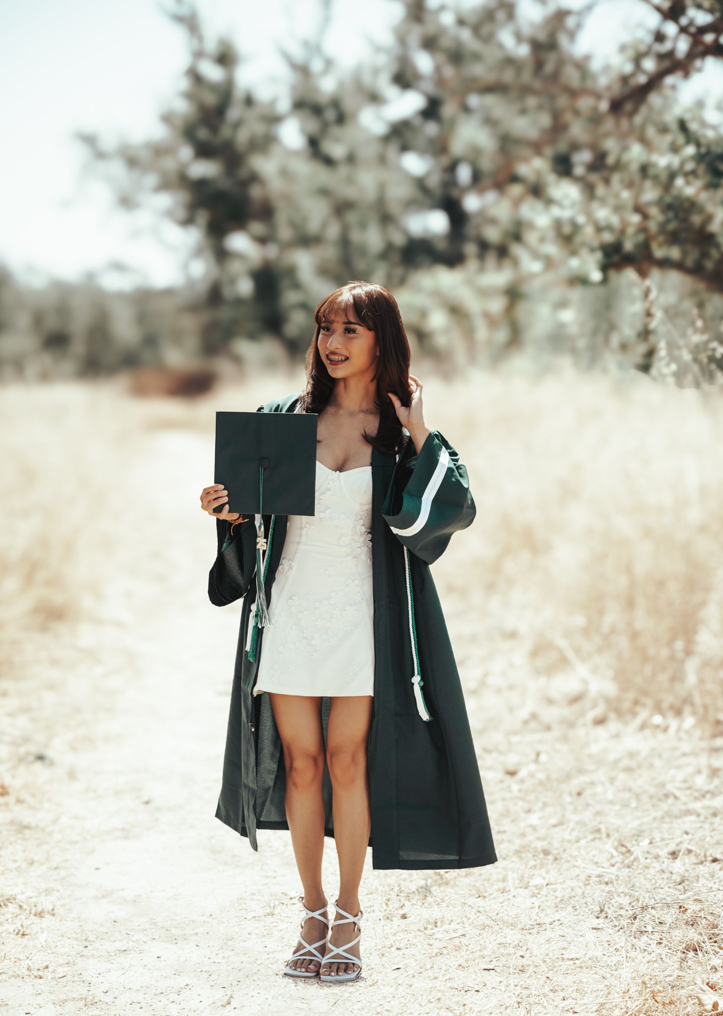A person in a white dress and long dark coat stands on a sunny path surrounded by blooming trees.