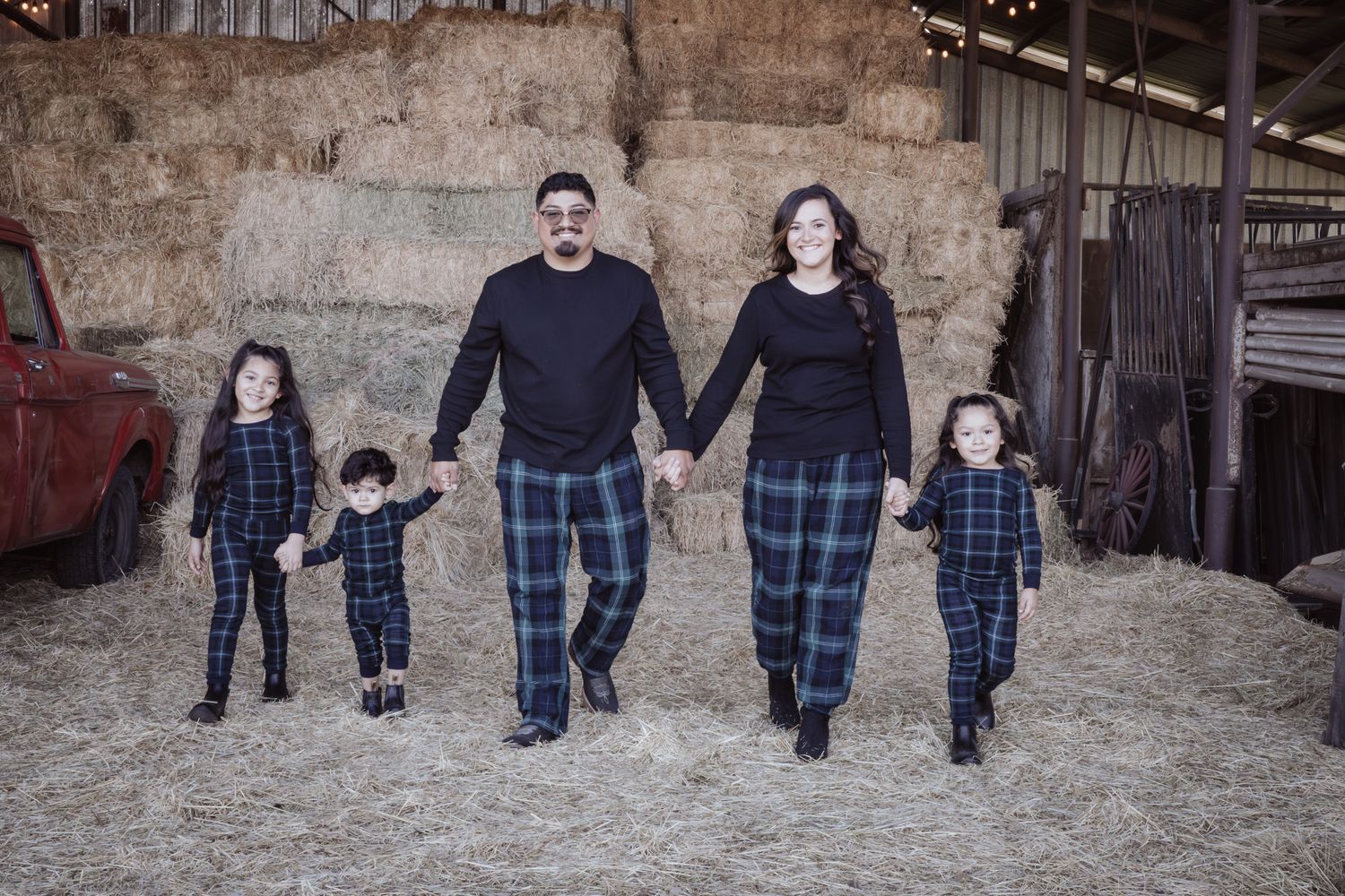 Family wearing coordinating plaid outfits walks together holding hands in front of rustic barn setting.