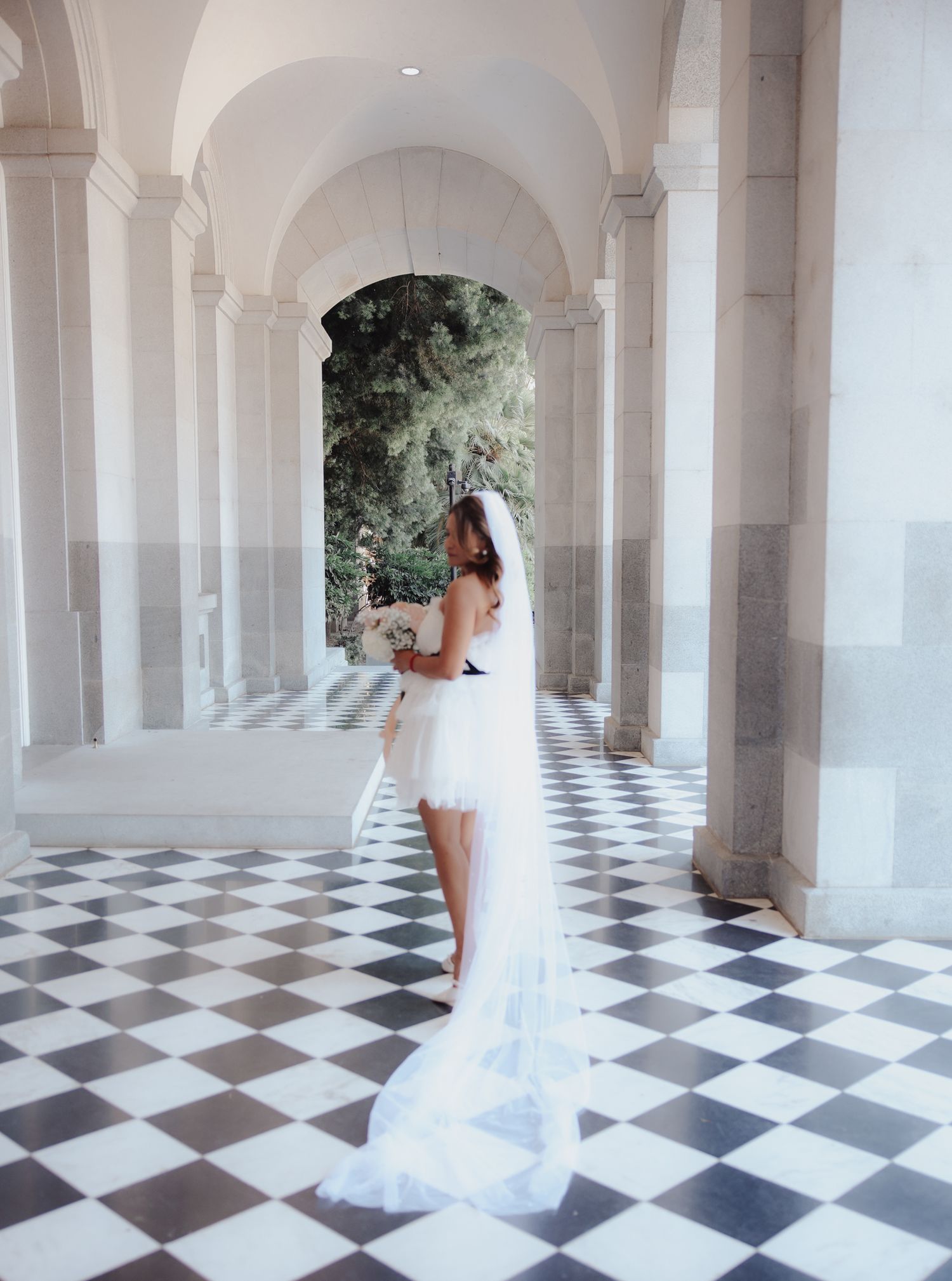 An elegant bride in a short white dress with flowing veil stands in a classical columned hallway with checkered floors.