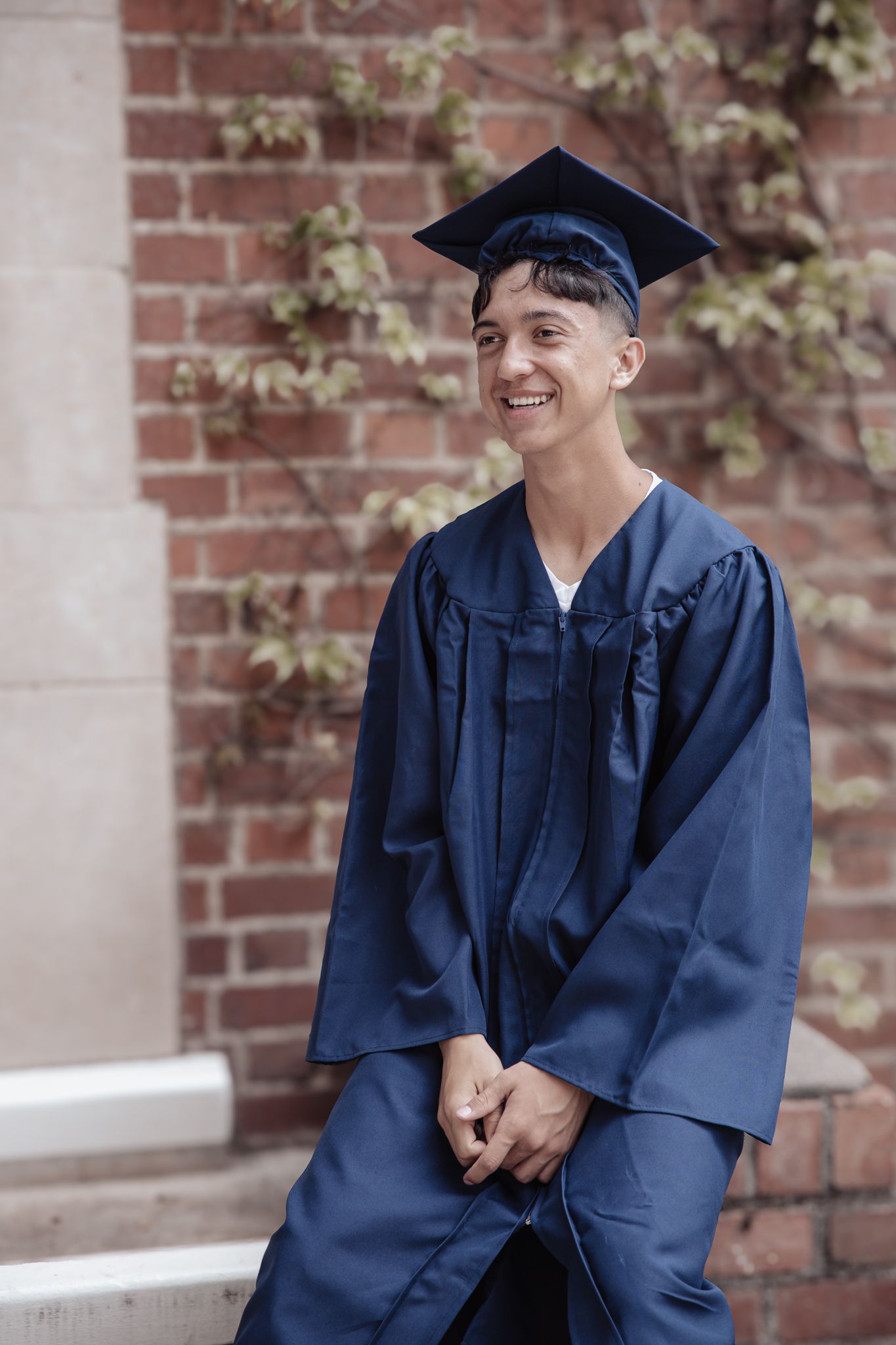 A graduate in a navy blue cap and gown smiles while standing against a brick wall.