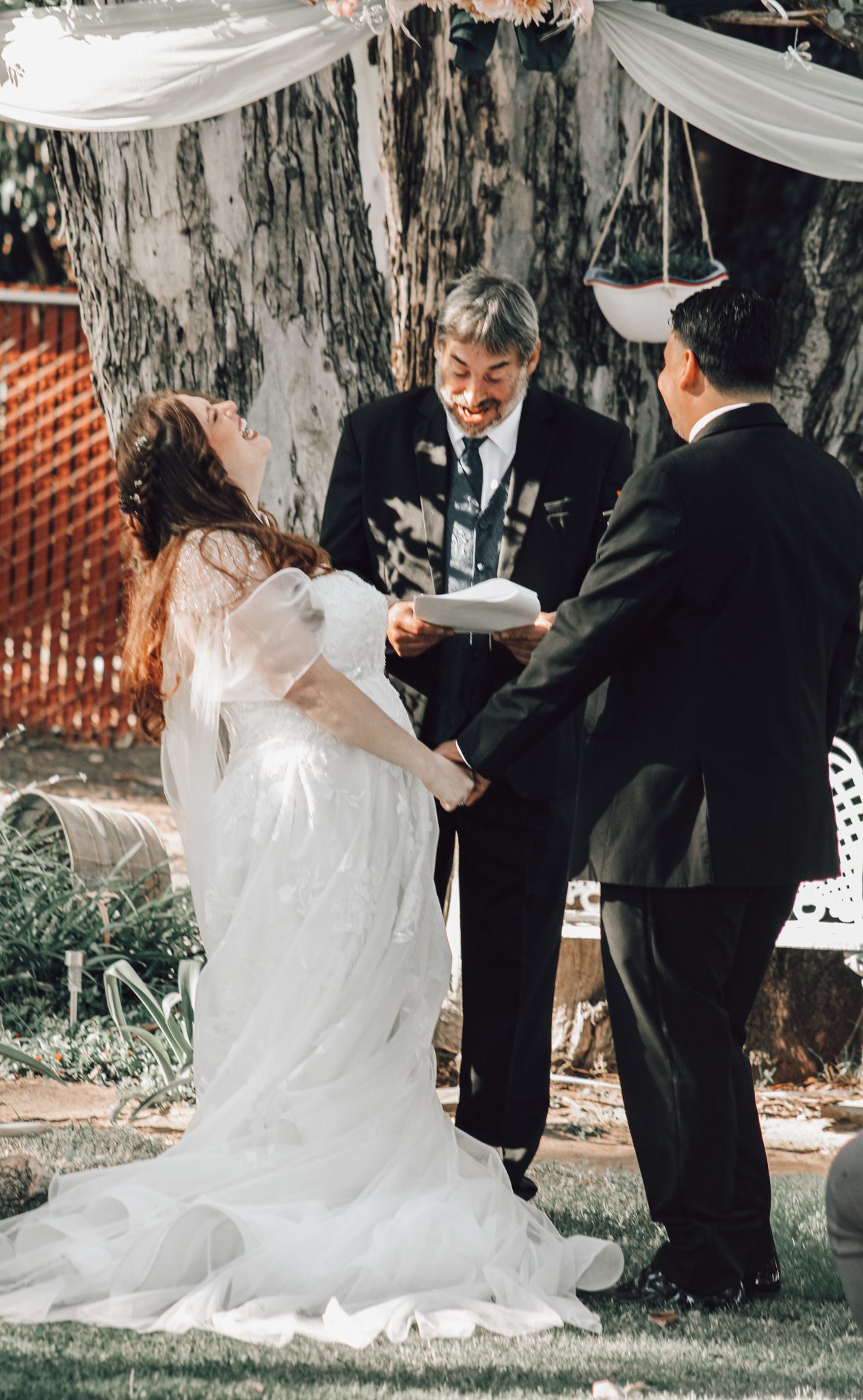 A couple in formal wedding attire shares a playful moment during their outdoor ceremony under draped white fabric.