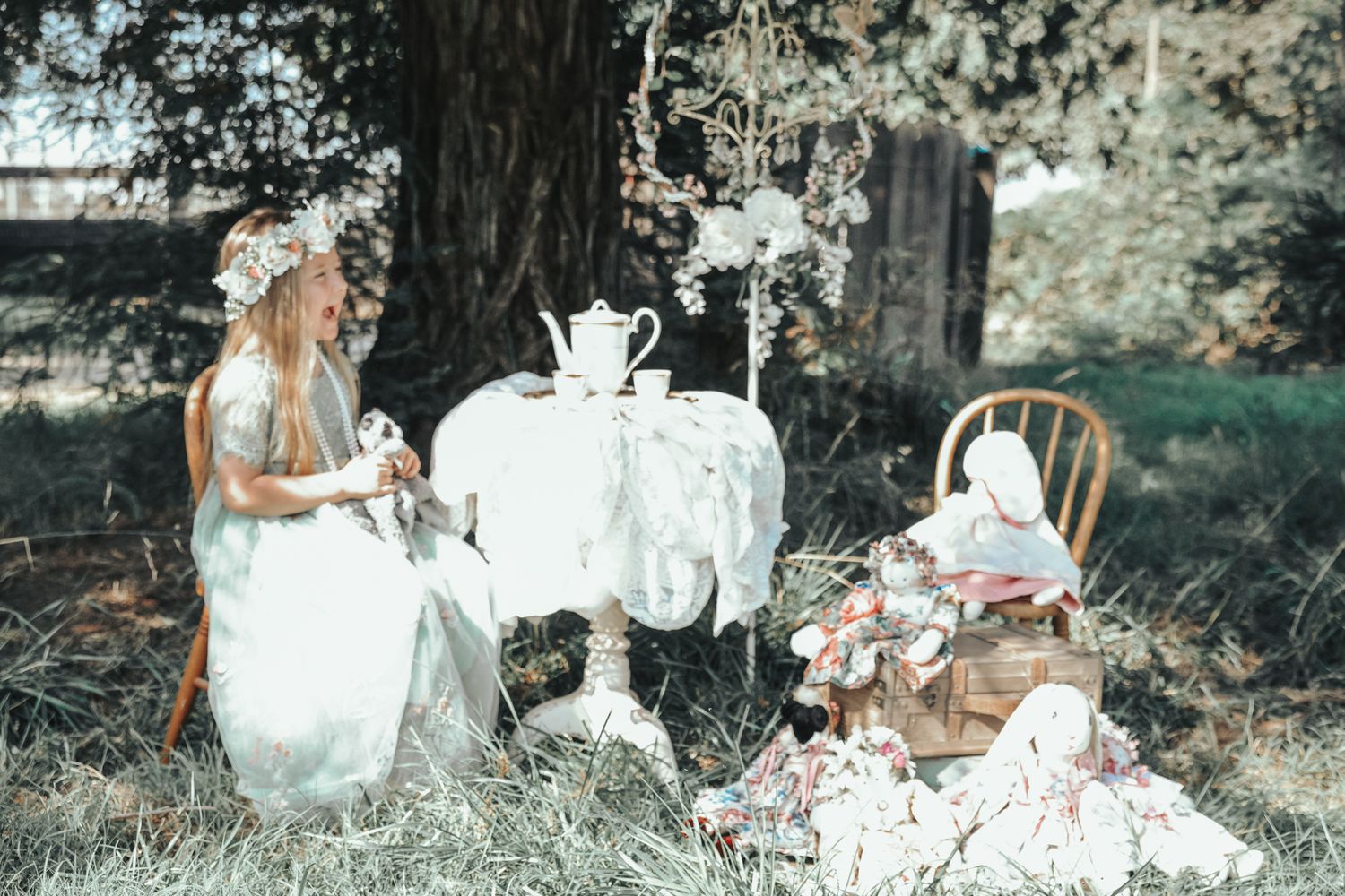 Girl in white dress with floral crown sits at outdoor tea party table under tree with doll in chair.