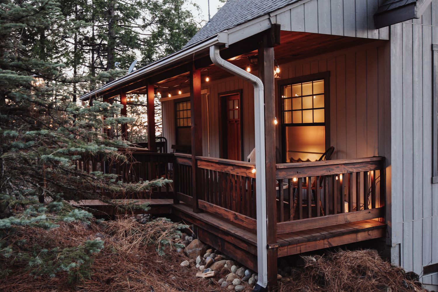 Cozy cabin porch with warm glowing lights and wooden railing nestled among evergreen trees at dusk.