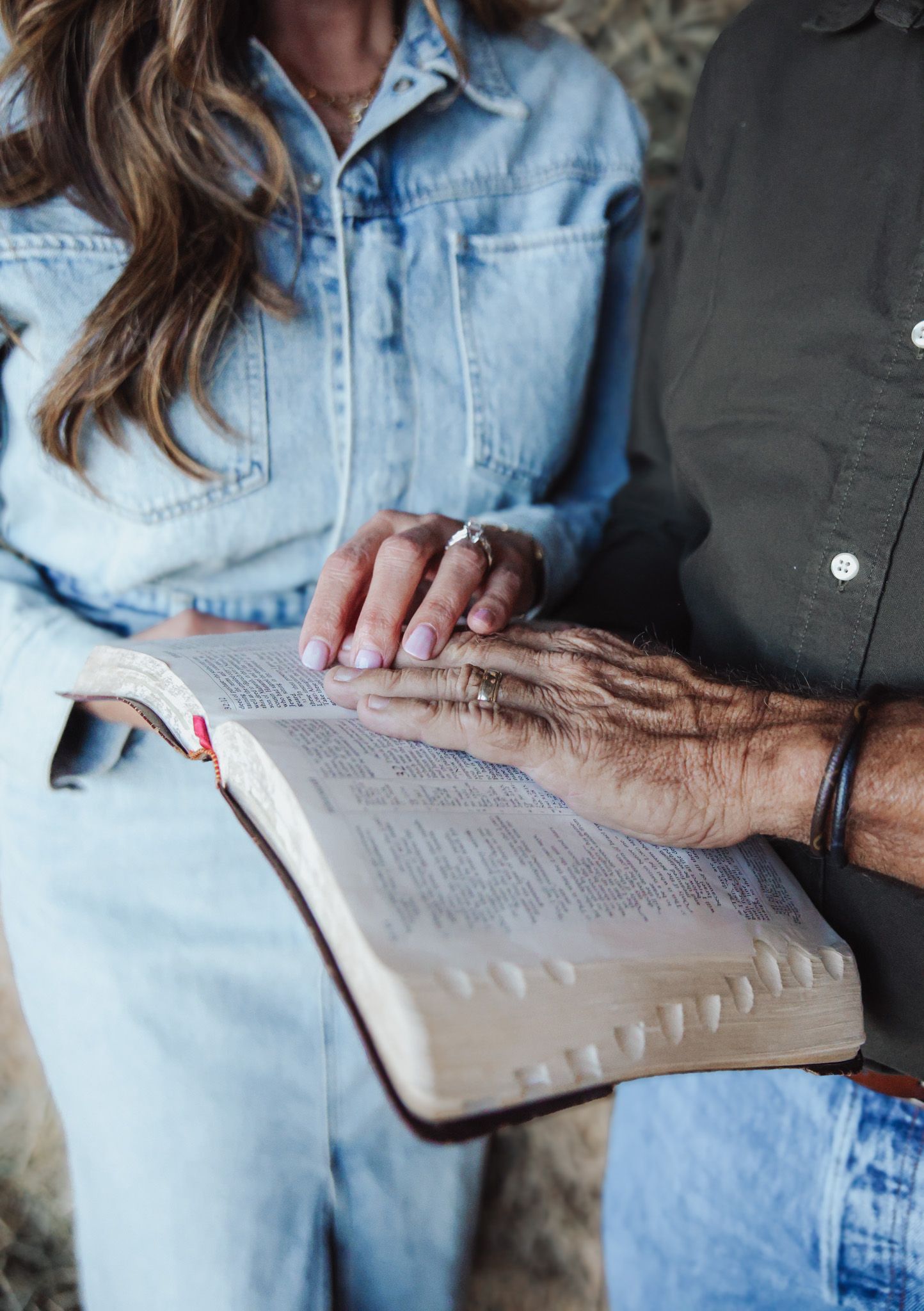 Person wearing denim shirt holds open Bible pages while studying scripture in natural lighting.
