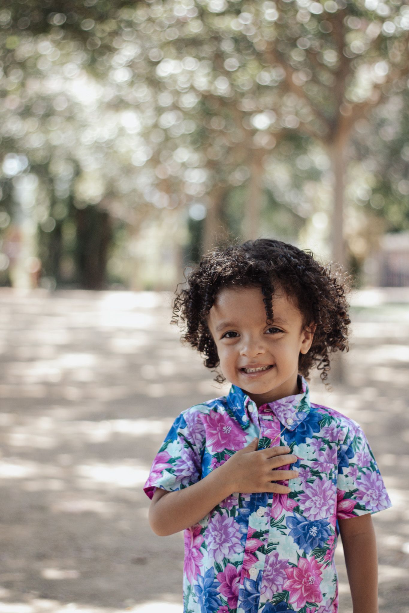 Young child wearing a colorful floral print shirt smiles while standing outdoors in dappled sunlight.