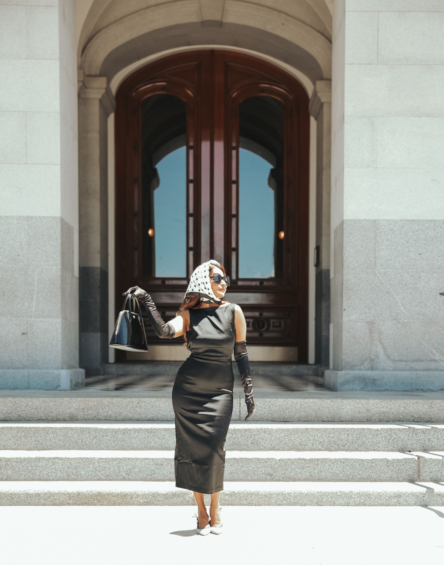 A woman in black dress walks away down a stone corridor with arched passageways and windows letting in natural light.