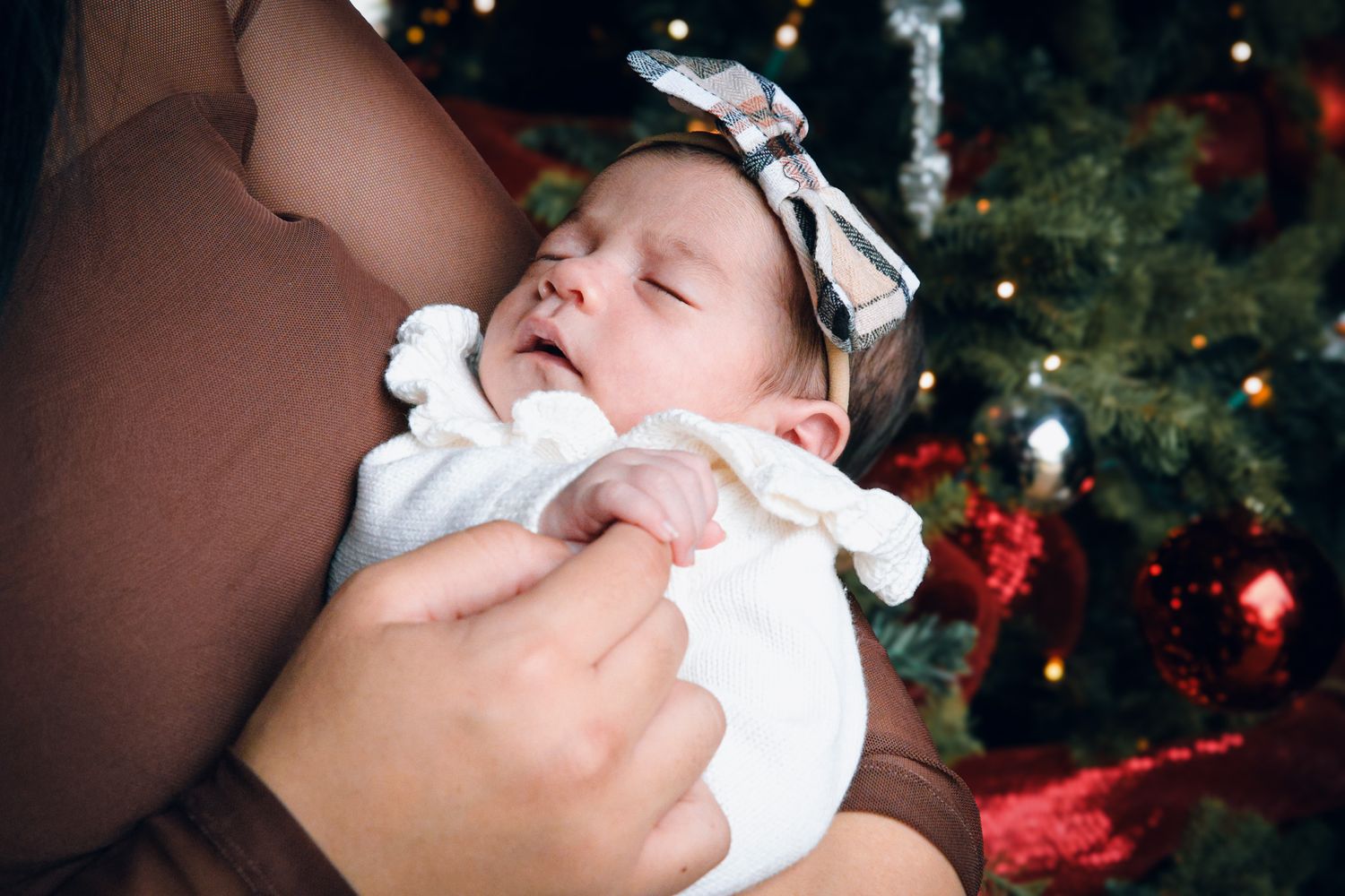 A sleeping baby wearing white clothing is cradled in someone's arms with a Christmas tree in the background.