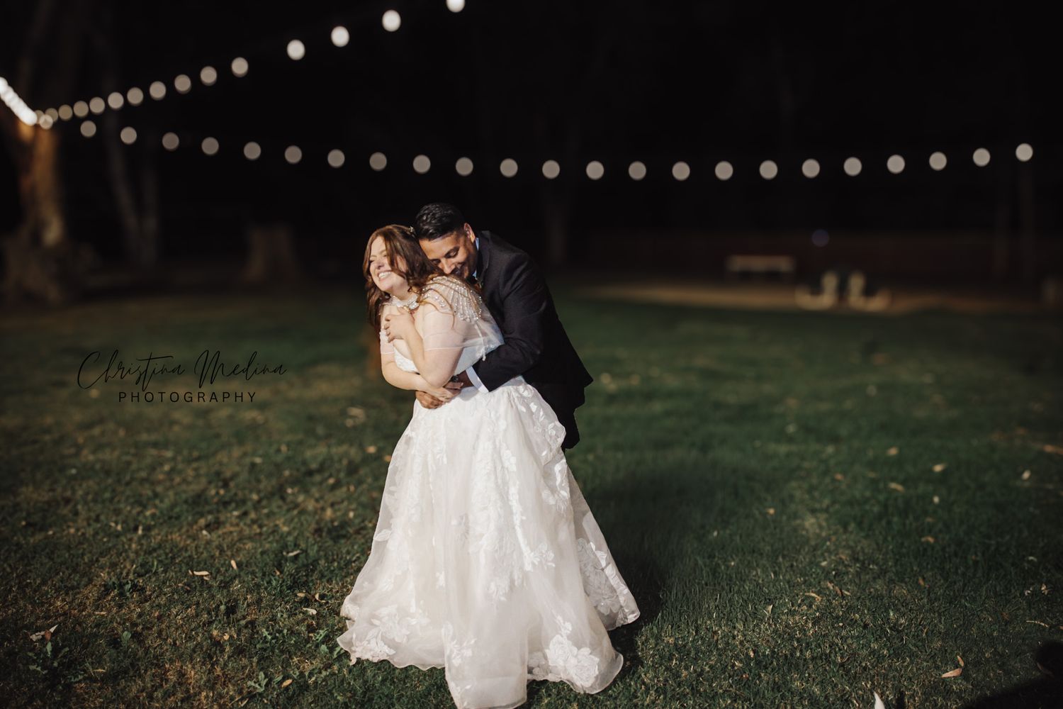 A couple shares a romantic moment under string lights on a dark grassy lawn during their wedding reception.