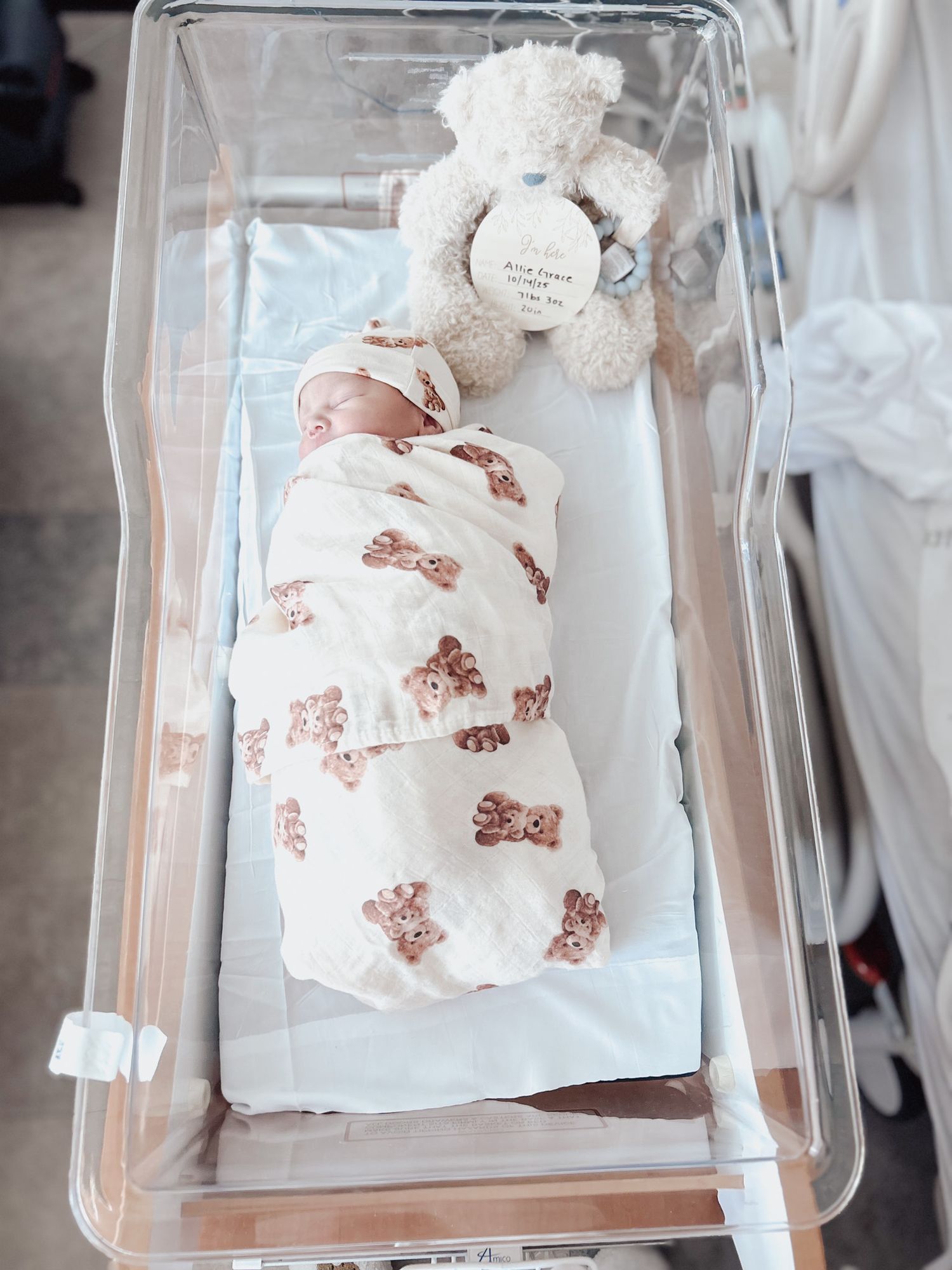 A newborn baby wearing a white outfit with teddy bear print lies in a clear hospital bassinet.