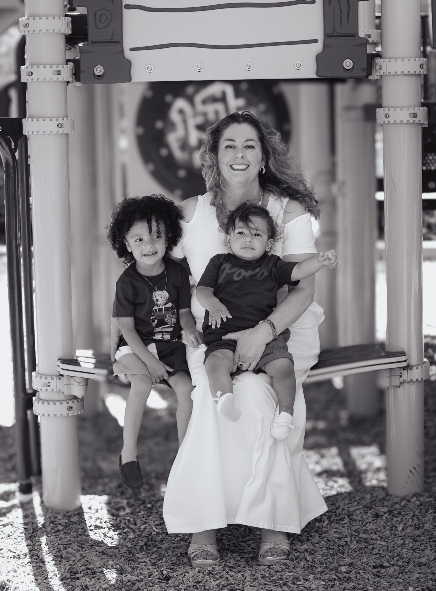 Black and white photo showing a joyful moment on playground equipment with two children and an adult in a white dress.
