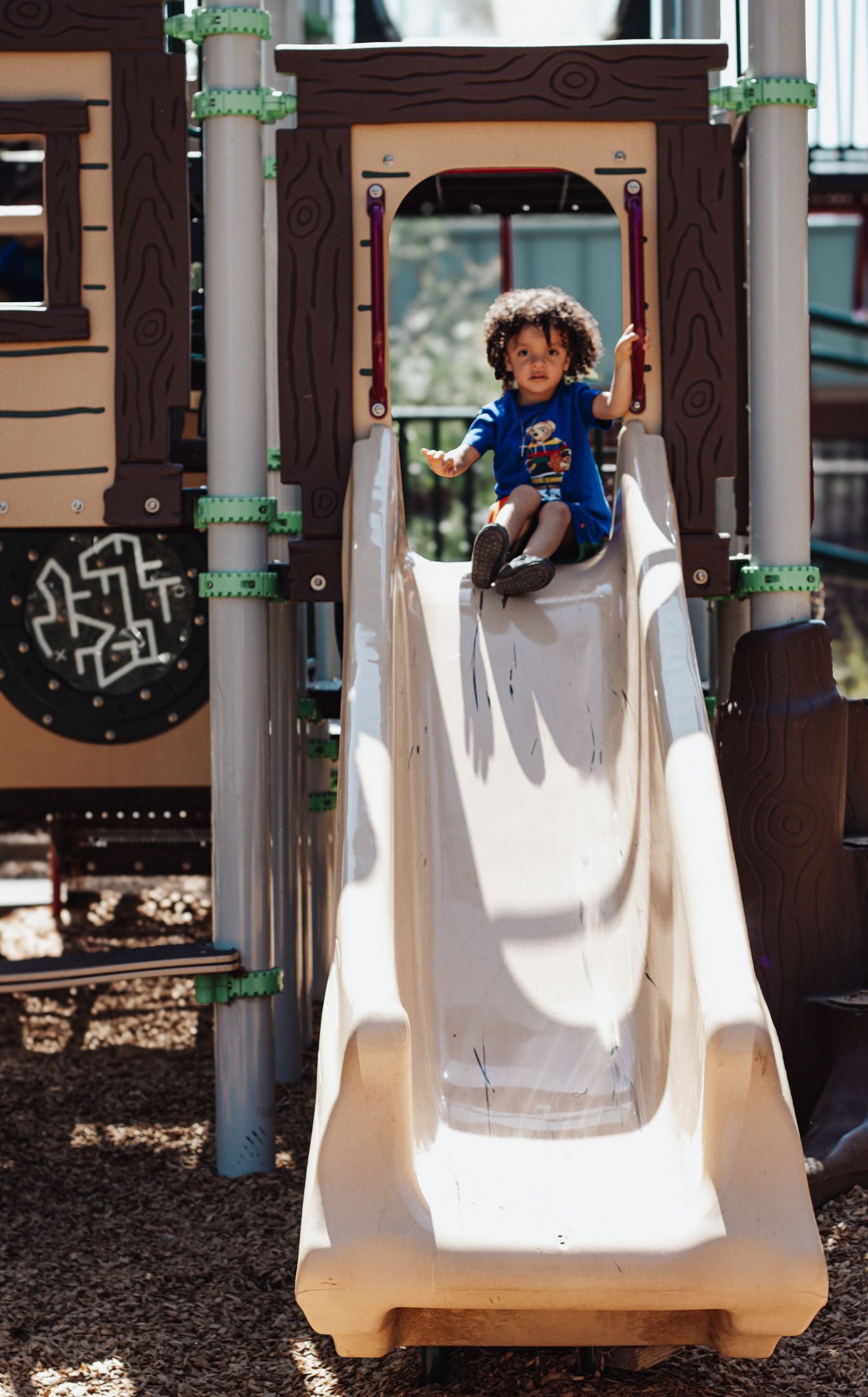 Young child in a blue t-shirt sits at the top of a white playground slide on a sunny day.