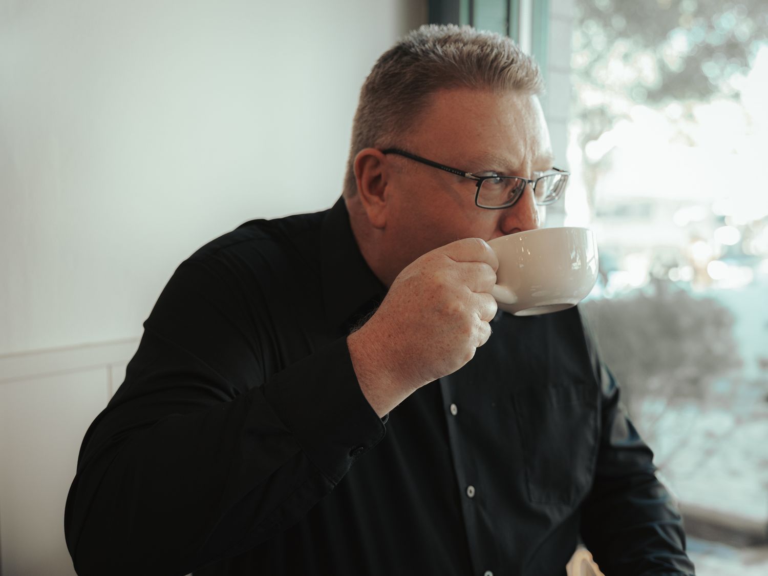 Someone in glasses and a black shirt sips from a white coffee cup while seated near a window.