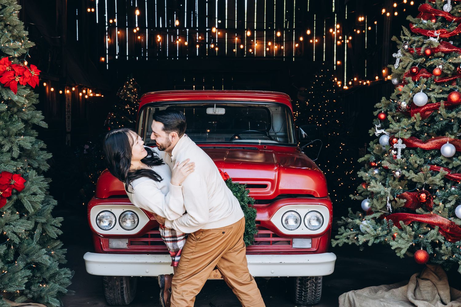 Couple in festive holiday attire poses romantically by vintage red car with Christmas decorations in background.