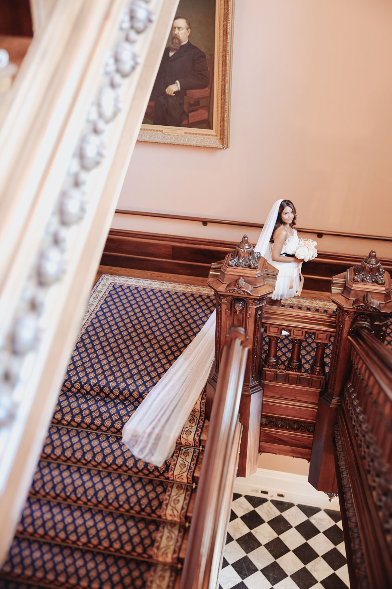 An elegant staircase with ornate wooden banisters, patterned carpet, and checkered marble floor in a grand Victorian interior.