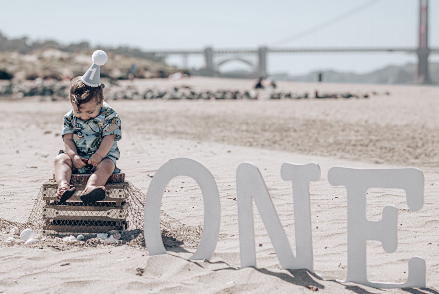 A young child sits on a rustic crate next to large white 'ONE' letters on a sandy beach with a bridge in the background.