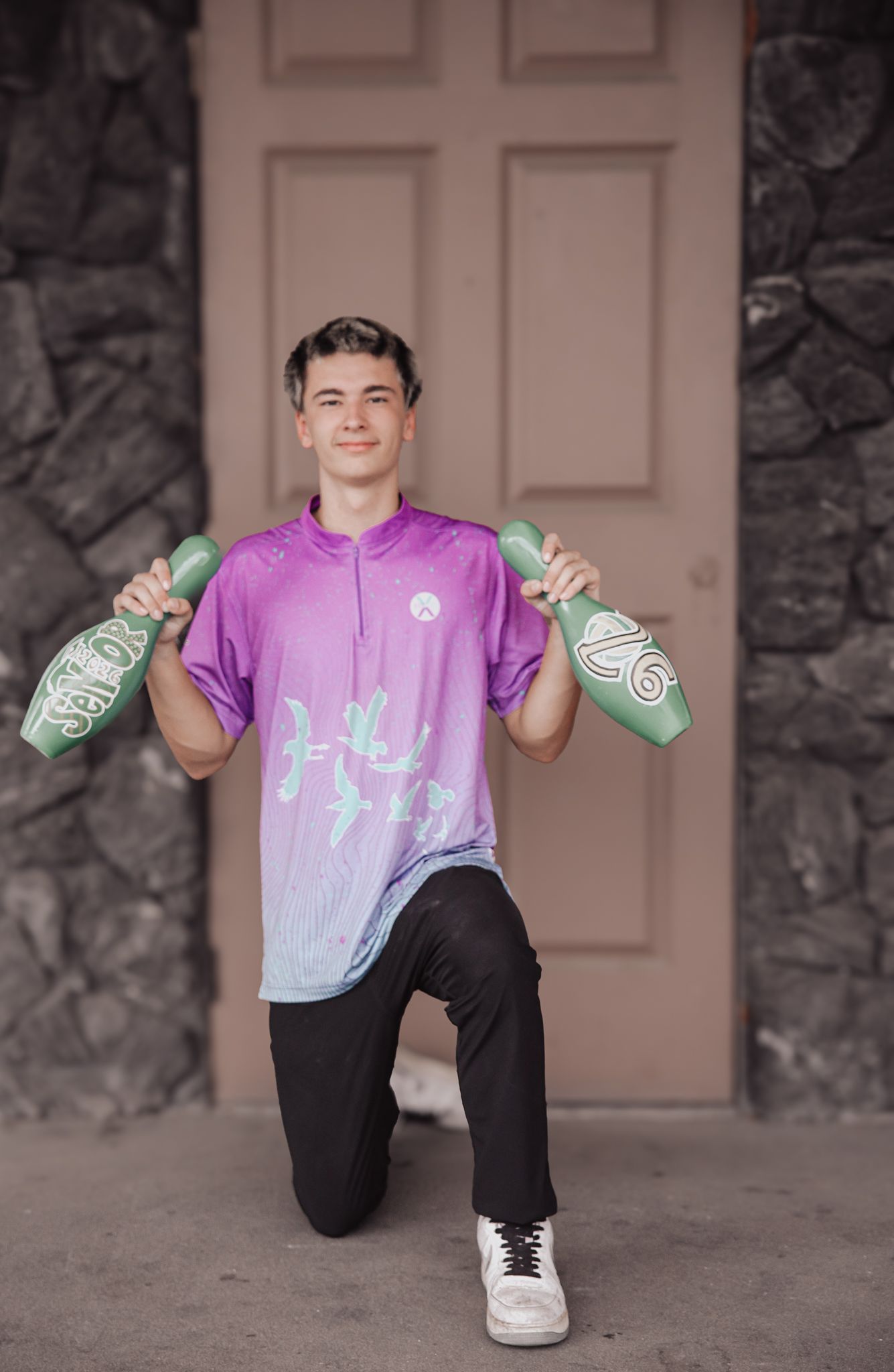 A person wearing a purple gradient shirt and black pants poses playfully on the ground with green gloves.