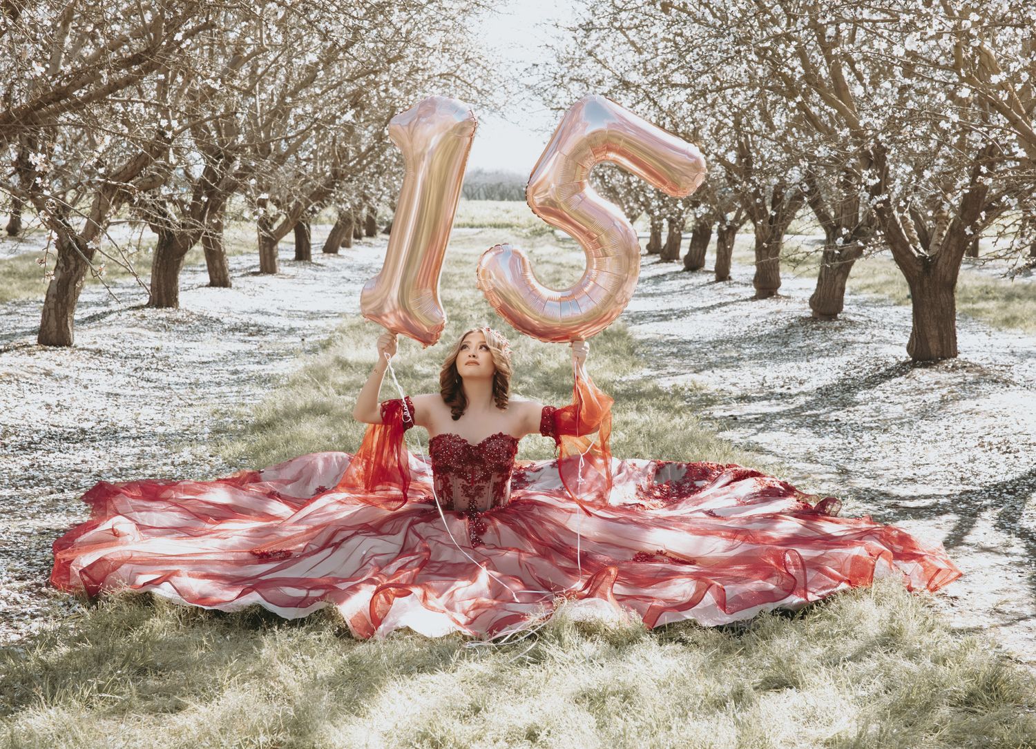 A red gown spreads across snowy ground between blooming almond trees with rose gold number 15 balloons floating above.