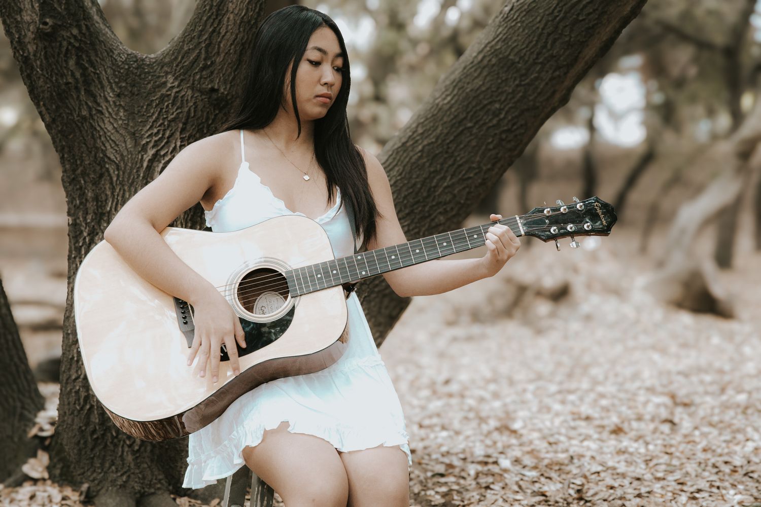 A person in a white dress plays acoustic guitar while sitting against a tree trunk in a dreamy outdoor setting.
