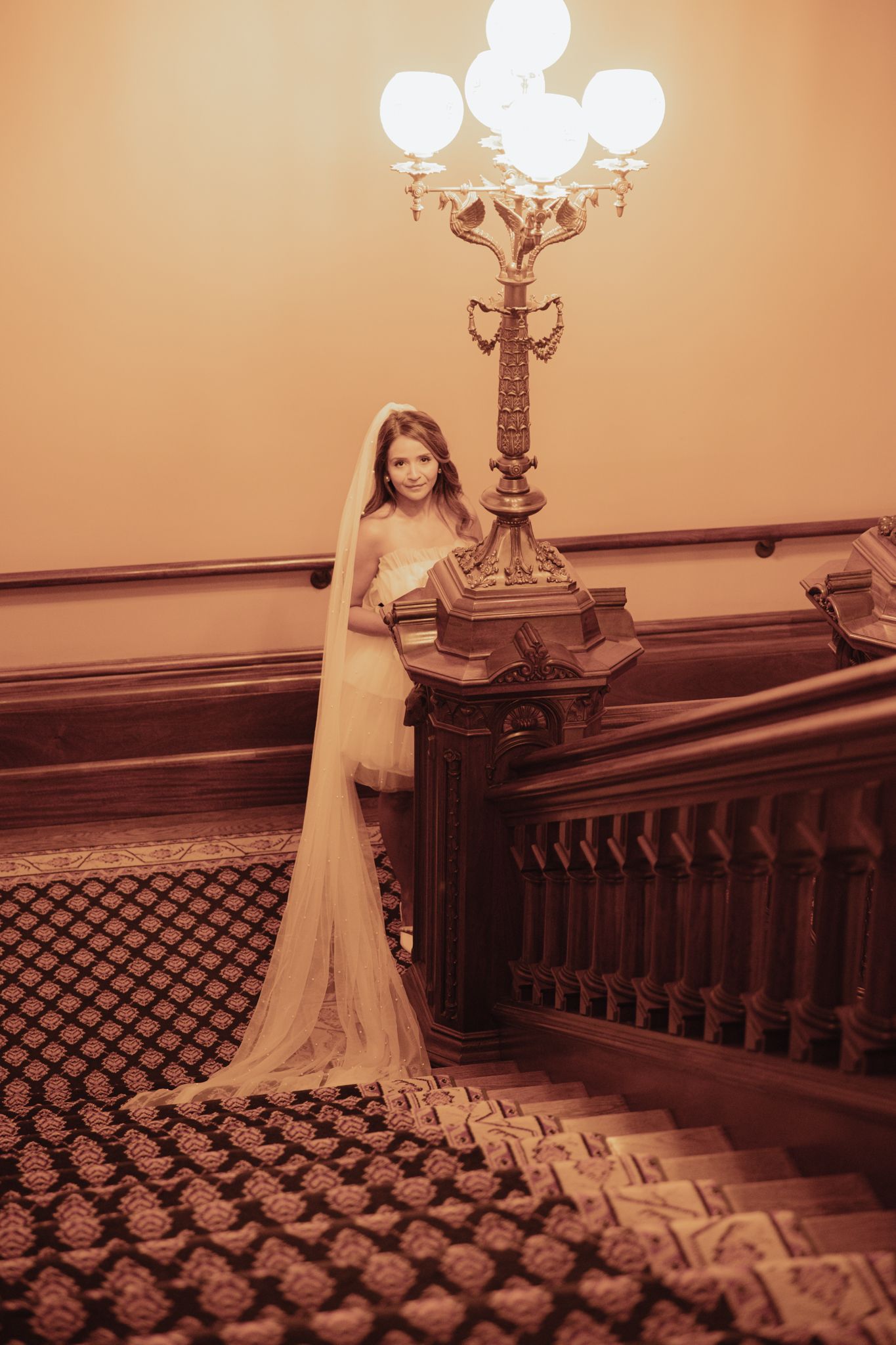 Figure in an elegant champagne gown poses on a grand staircase with ornate lighting and carpet in sepia tones.