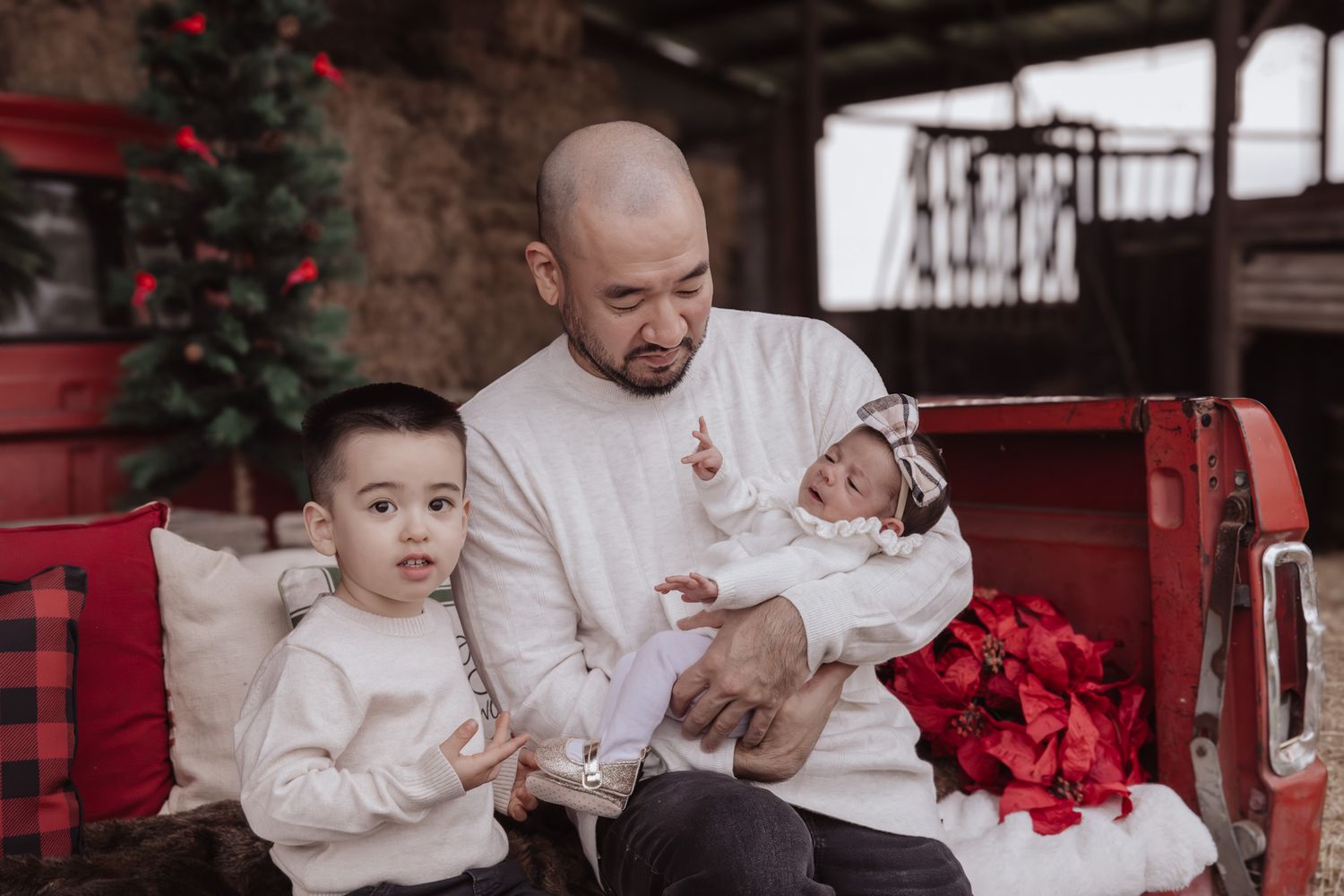 A family in white outfits sits together for a holiday photo session with red poinsettias and rustic decor.