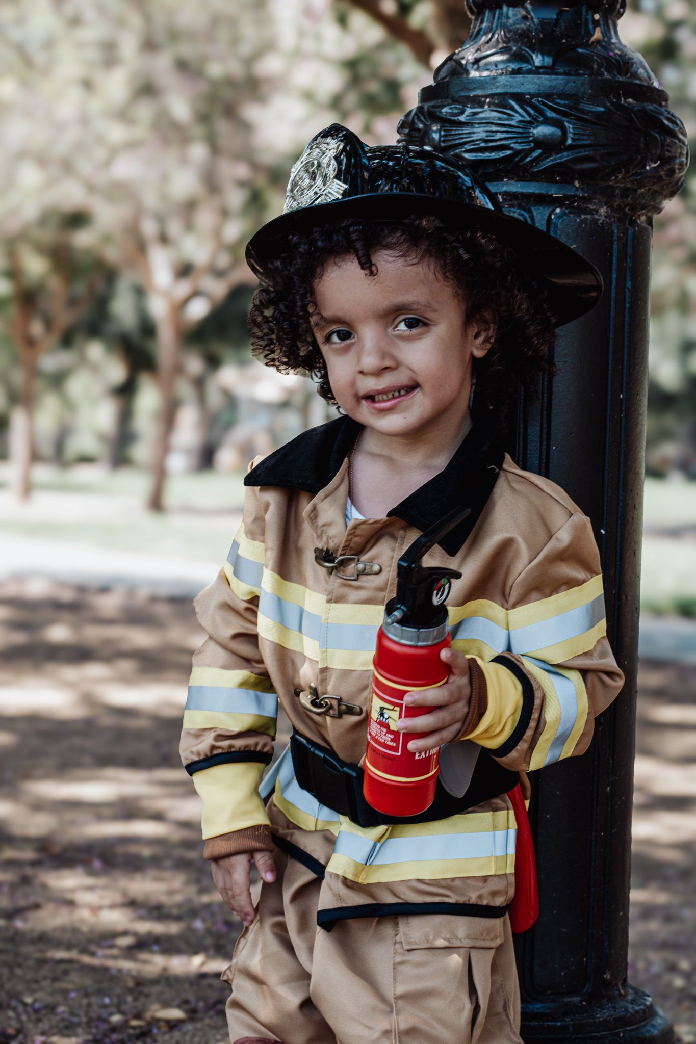 A young child wearing a firefighter costume and holding a red water bottle stands next to a black lamp post in a park.