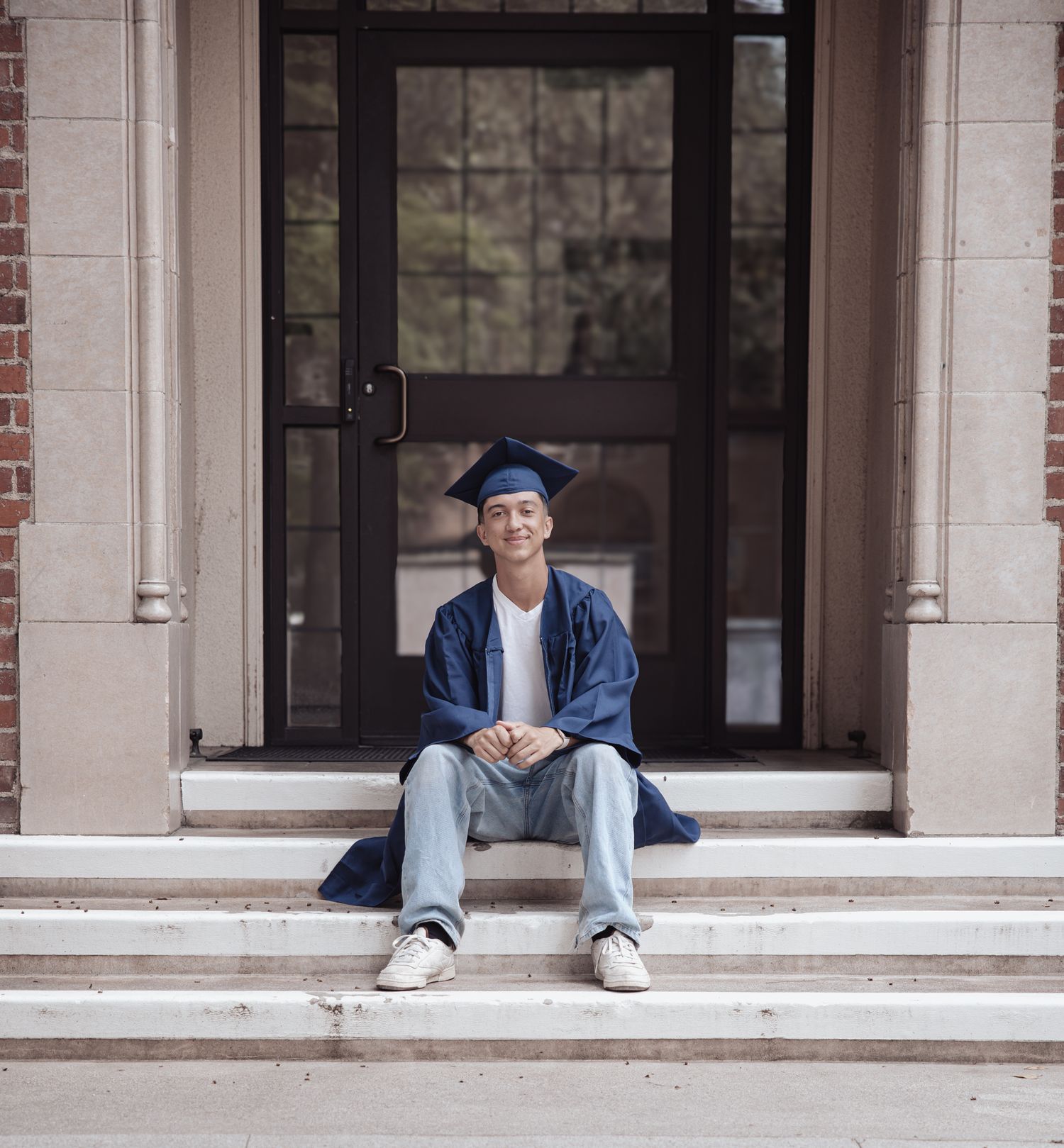 A person in graduation attire sits on outdoor stone steps in front of a dark glass door entrance.