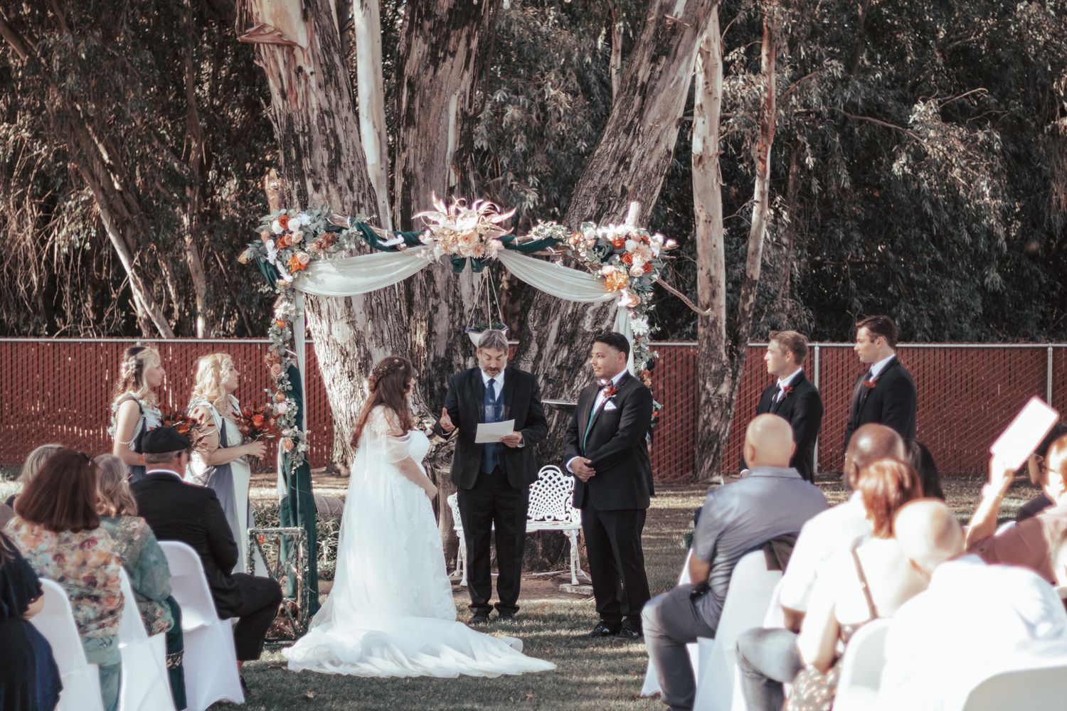 An outdoor wedding ceremony takes place under a floral arch with eucalyptus trees and guests seated on white chairs.