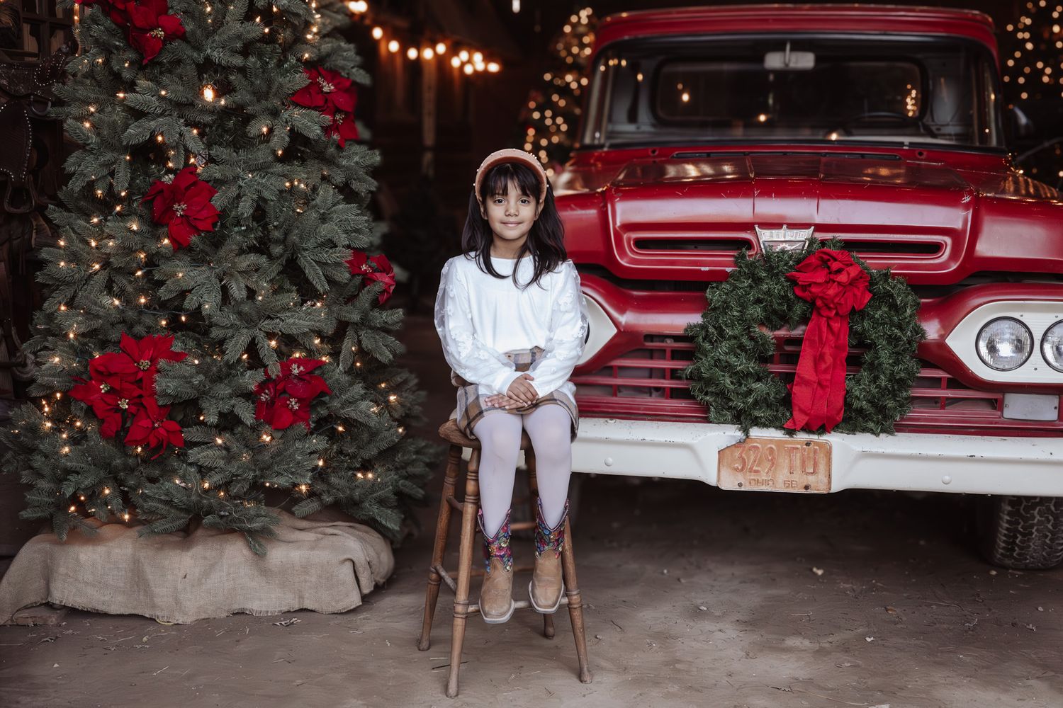 A vintage red pickup truck decorated with Christmas lights parked in front of an evergreen tree with holiday decorations.