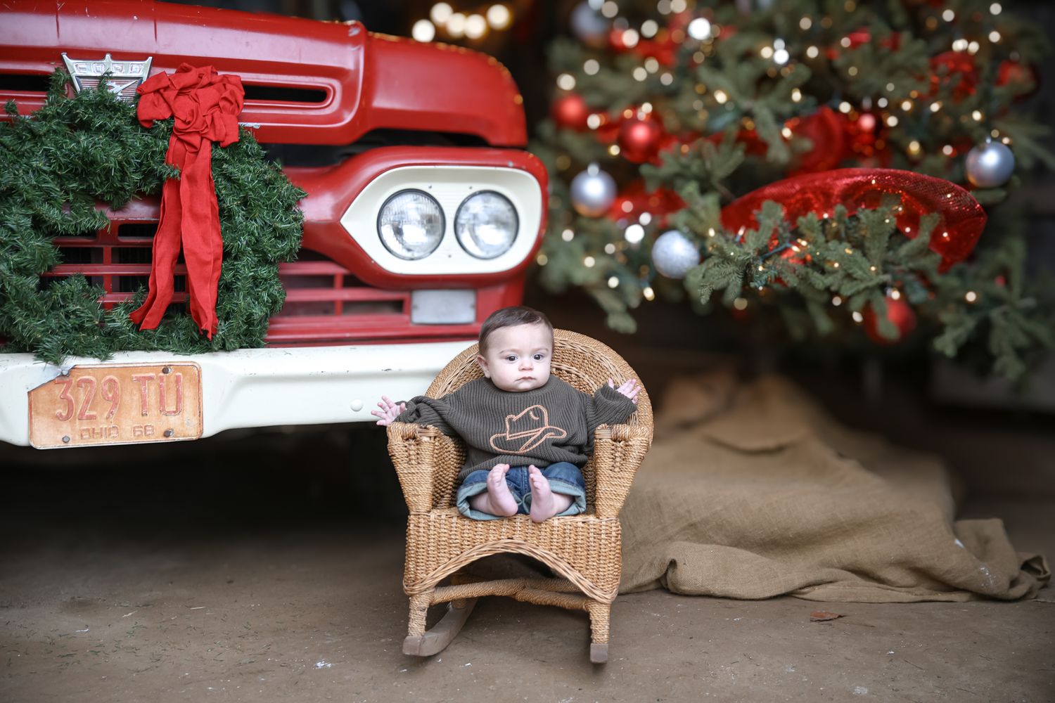 Vintage red truck with Christmas wreath and wicker chair displayed in front of decorated Christmas tree.