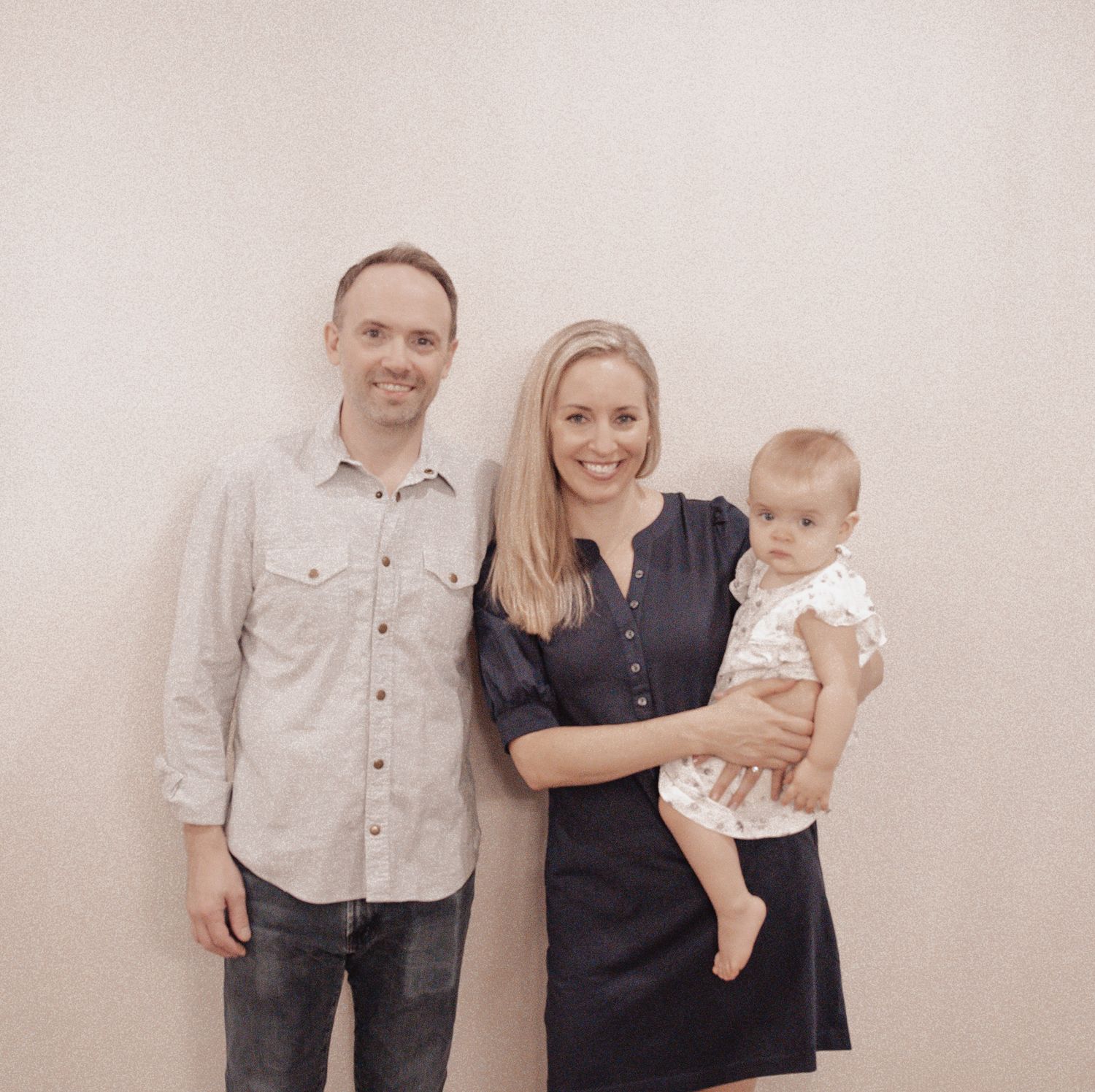 A family of three poses together against a light-colored wall in matching neutral-toned outfits.