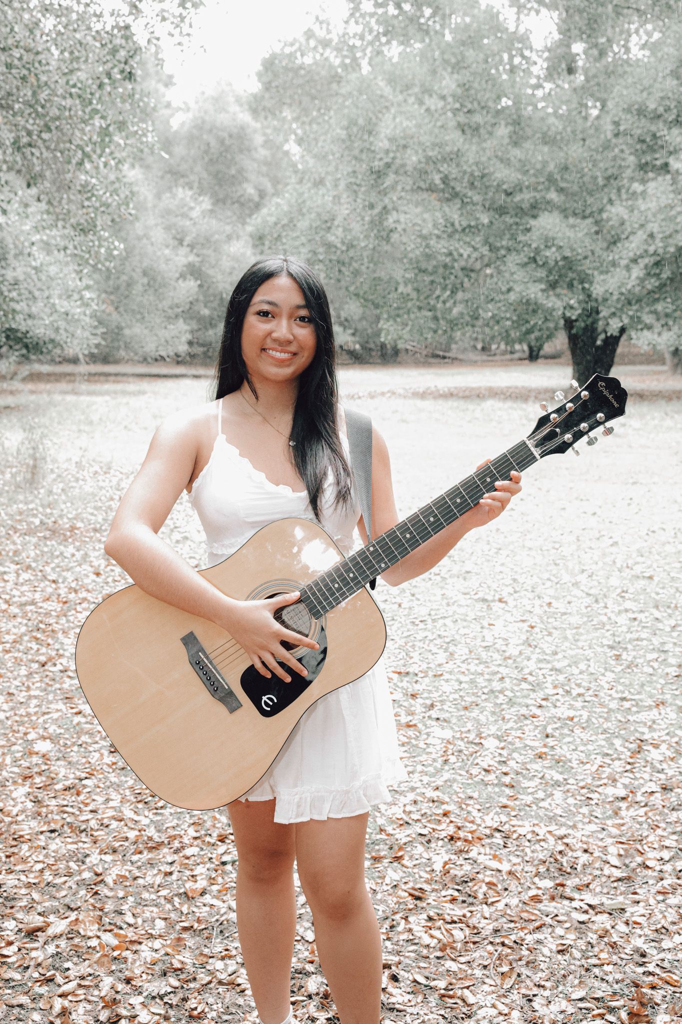 A musician in a white dress plays an acoustic guitar while standing in a park with fallen autumn leaves.