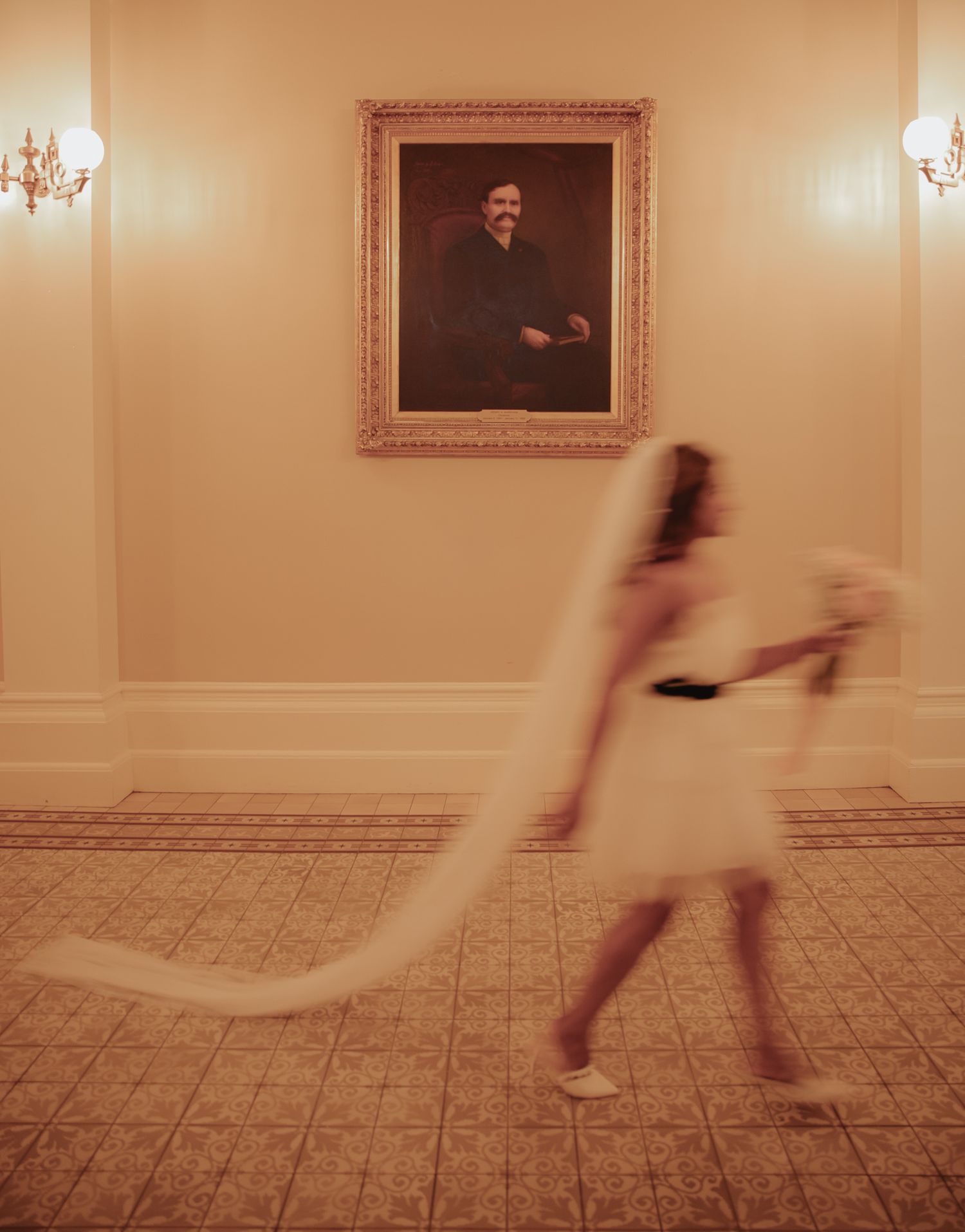 A motion-blurred figure in white walks past an ornate portrait and wall sconces in an elegant hallway.
