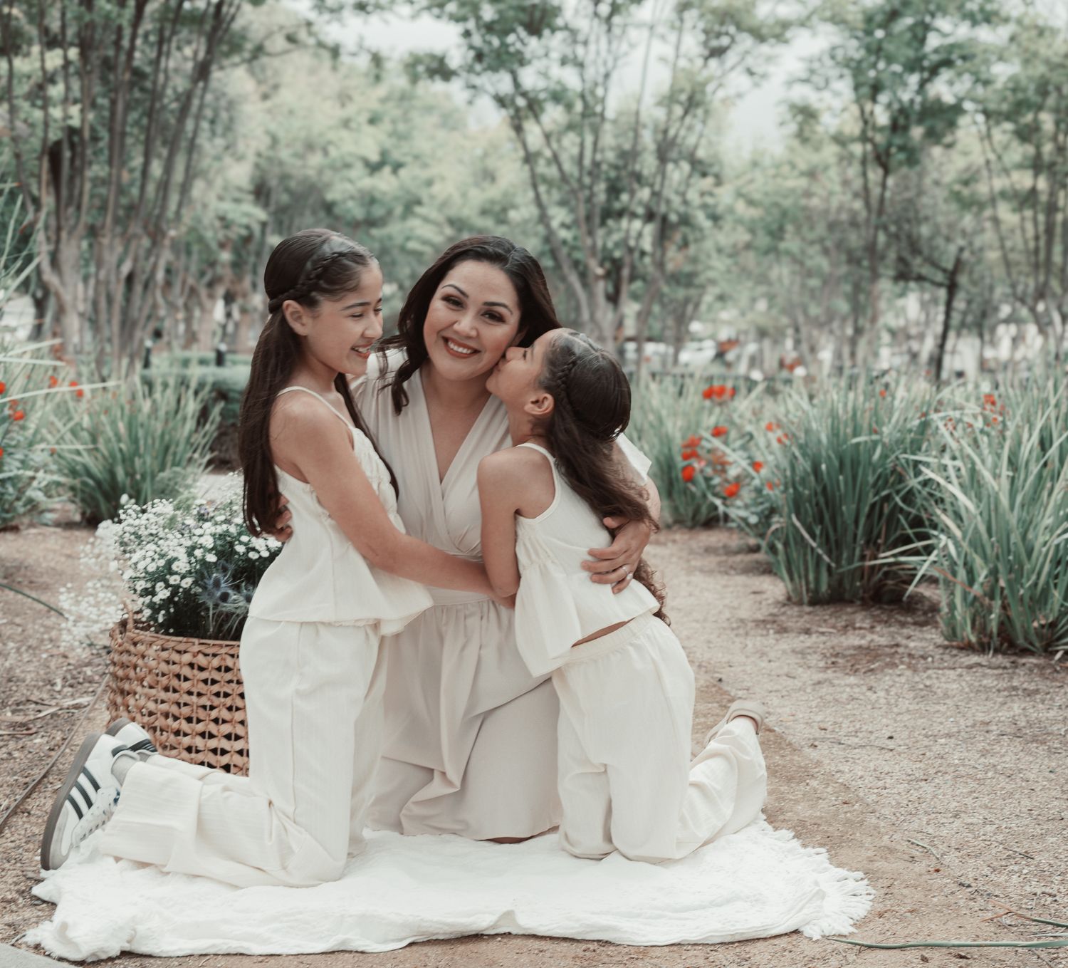 Three people in white outfits share a tender moment while sitting on a blanket in a lush garden setting.