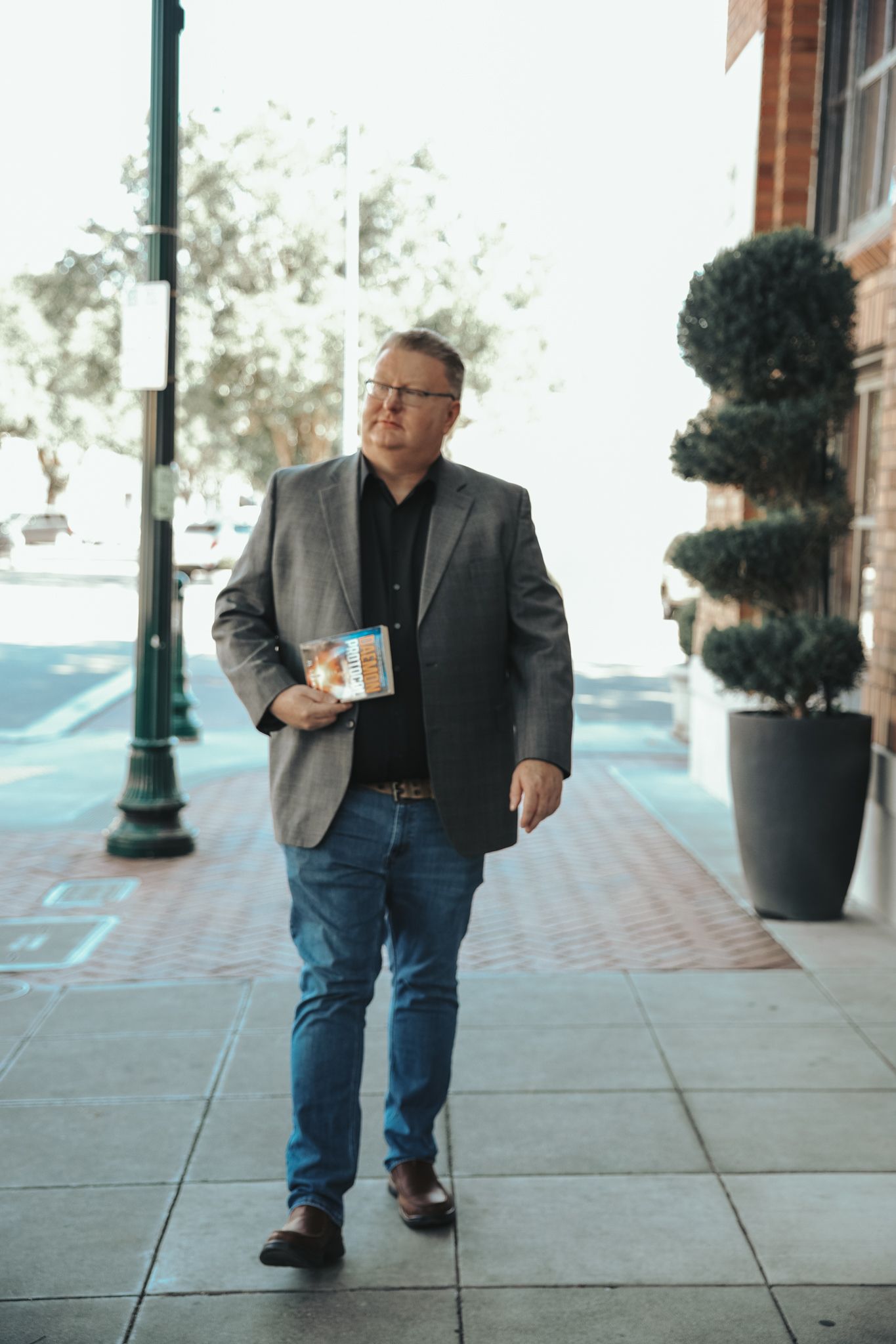 A person in a gray blazer and jeans walks along a sidewalk with decorative planters and lamp posts.
