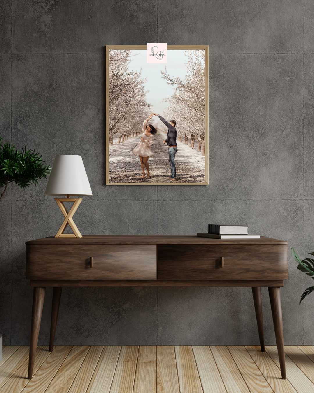 Modern wooden console table with crossed legs and lamp against concrete wall with framed artwork in contemporary setting.