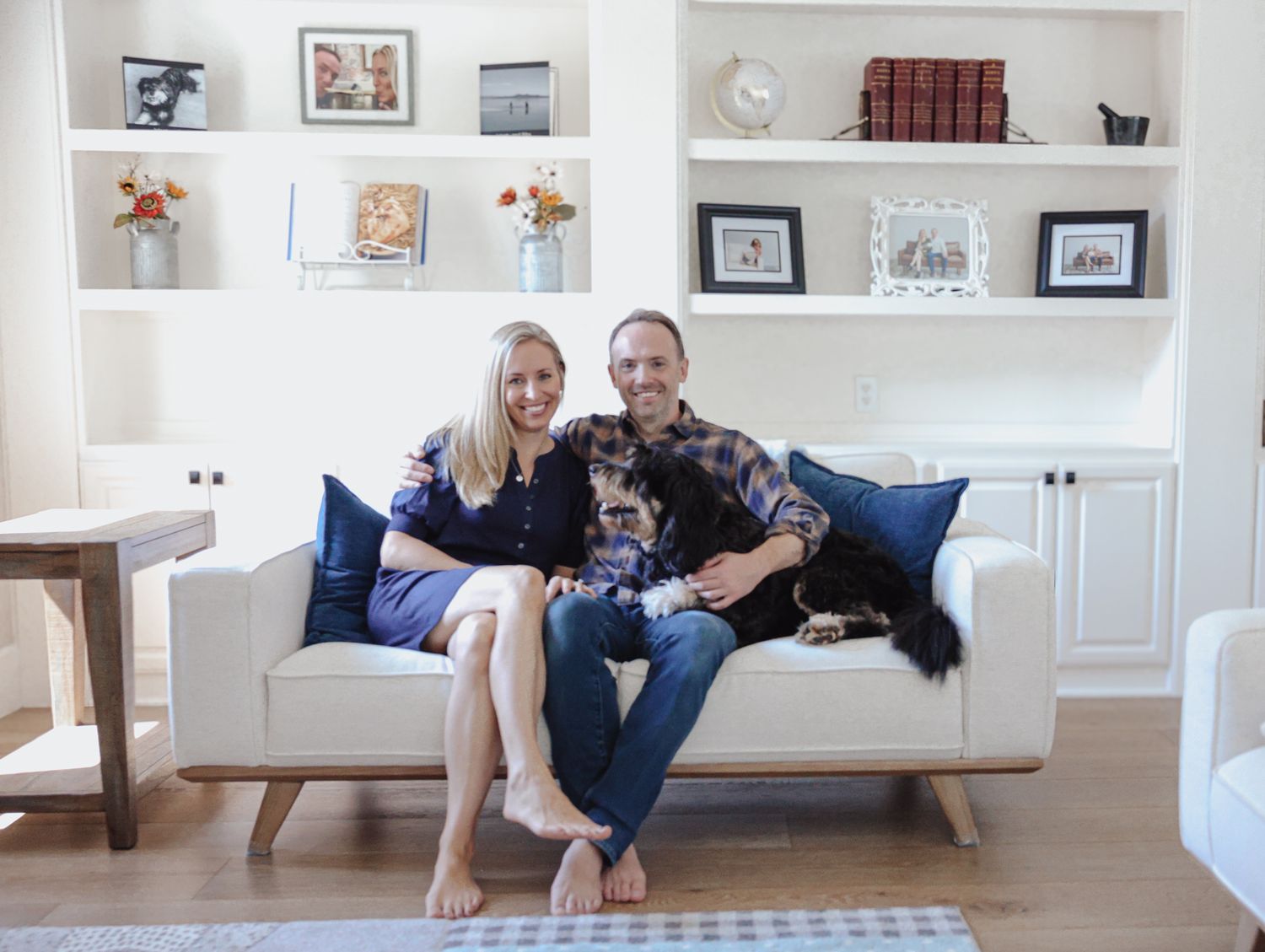 A couple relaxes on a white couch with two dogs in a bright living room decorated with floating shelves and blue pillows.