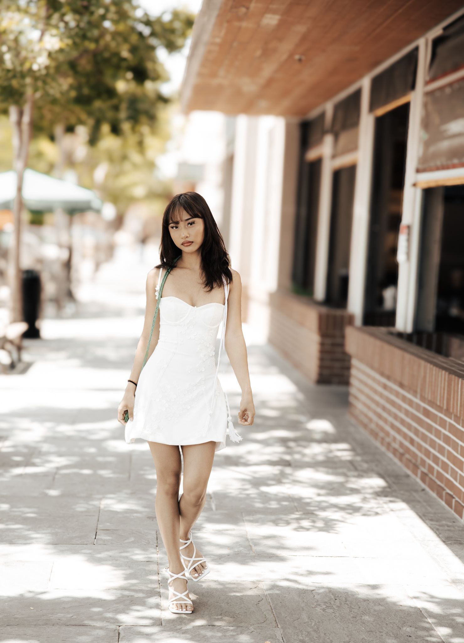 Person in a short white summer dress and sandals walks along a sunny sidewalk near a brick building.