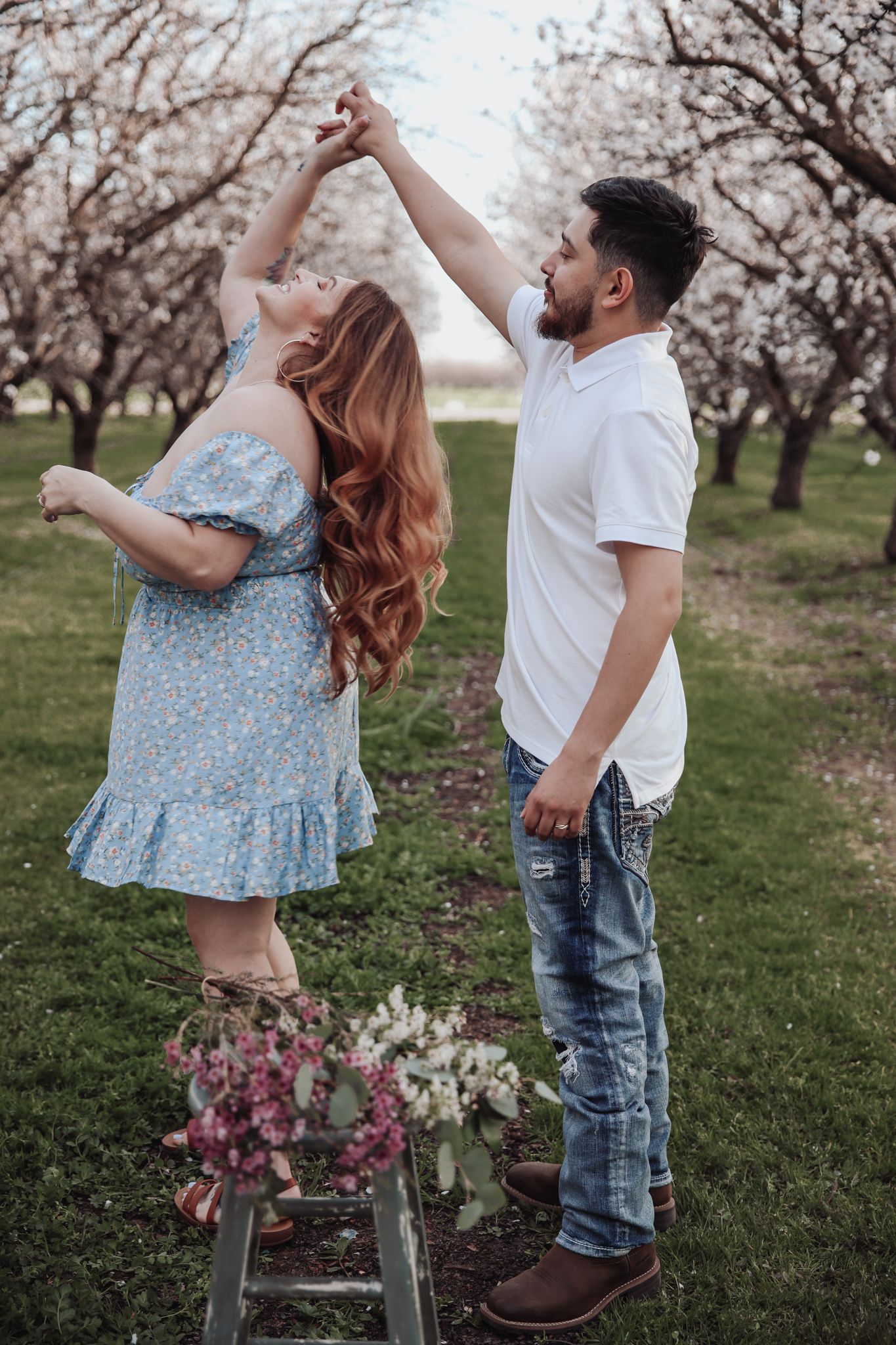 A couple dances together in a blooming almond orchard during springtime wearing casual attire.
